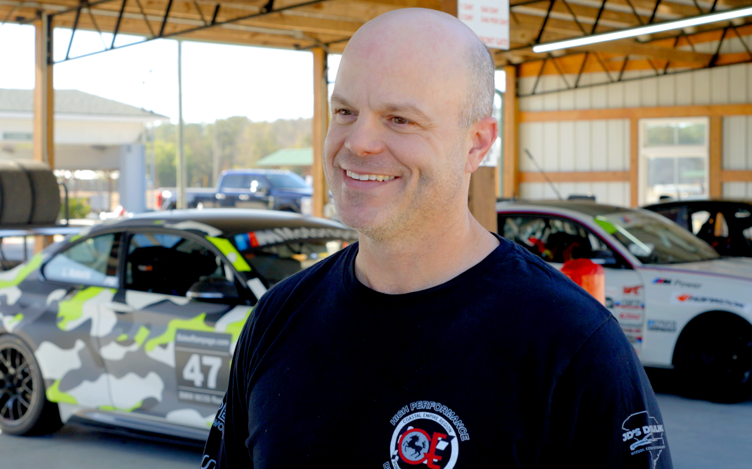 Co-Founder, Rich Exline, smiling in a black race team shirt standing in a garage with race cars behind him.