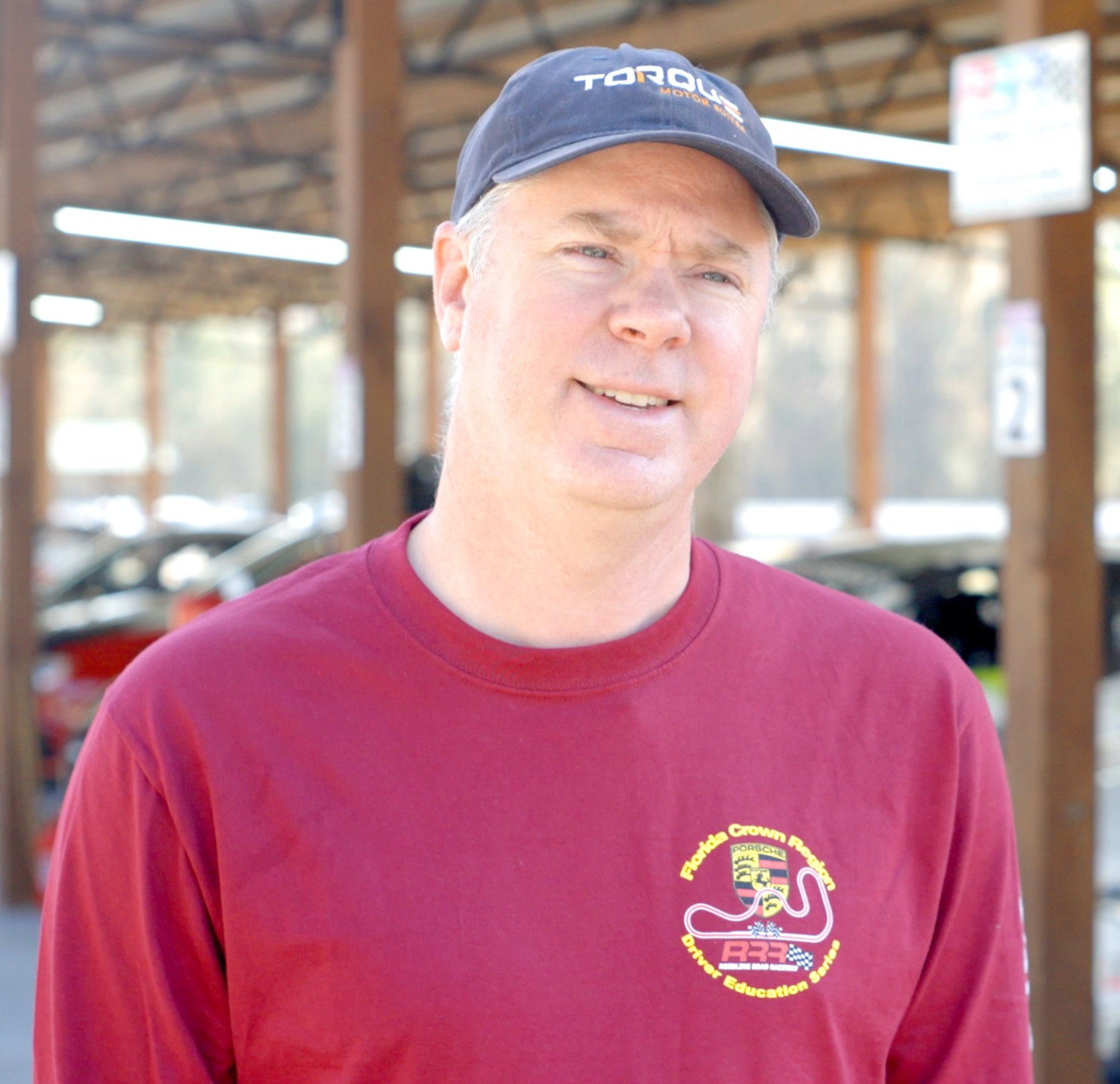 Co-Founder, Charles Roberts, wearing a navy baseball cap with the word 'TORQUE' printed on it, a maroon long sleeve shirt with a Florida Crown Racing logo, and standing outdoors in a parking lot with cars and a wooden shelter in the background.