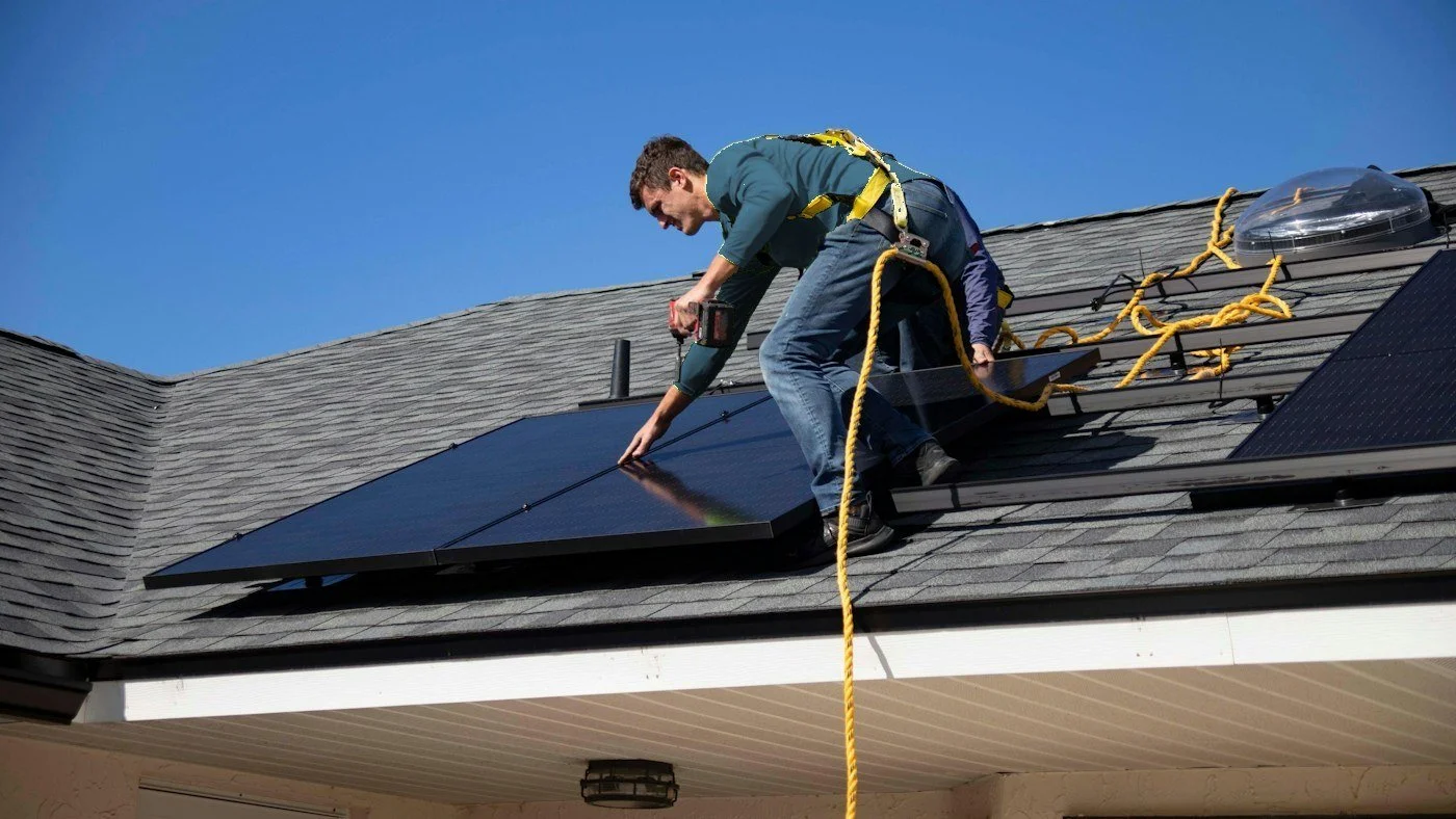Technician in a safety harness working on rooftop solar panels on a shingled home