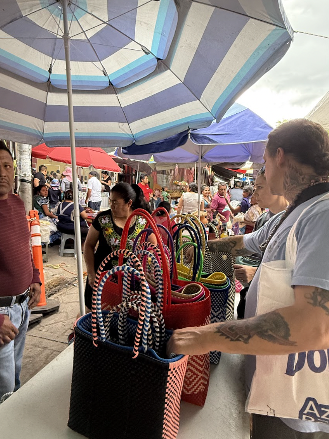 A bustling outdoor market with colorful umbrellas overhead, selling various woven bags. Several people are shopping, browsing, and engaging with vendors under a cloudy sky.