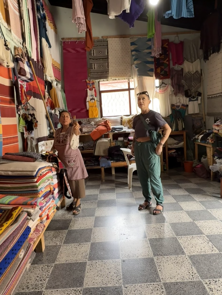 Two women standing inside a textile shop with colorful fabrics on shelves and hanging from walls and ceiling, with a window in the background.