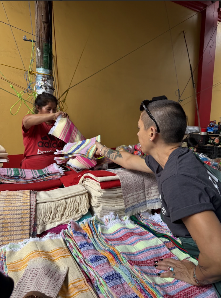 A woman in a red shirt buys colorful woven hand towels from a vendor at a market stall.