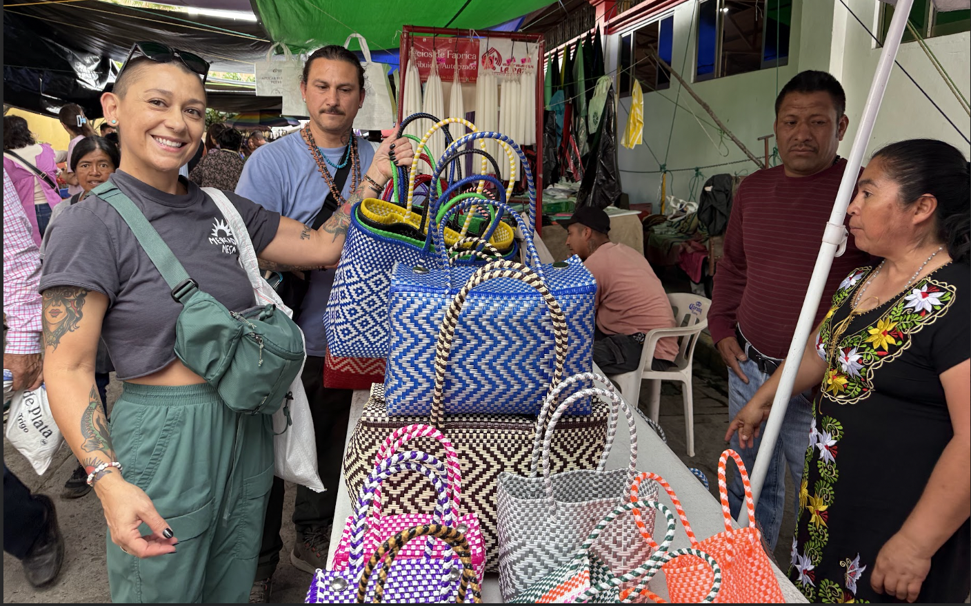 People at a market stall display colorful woven bags, with a woman smiling at the camera.