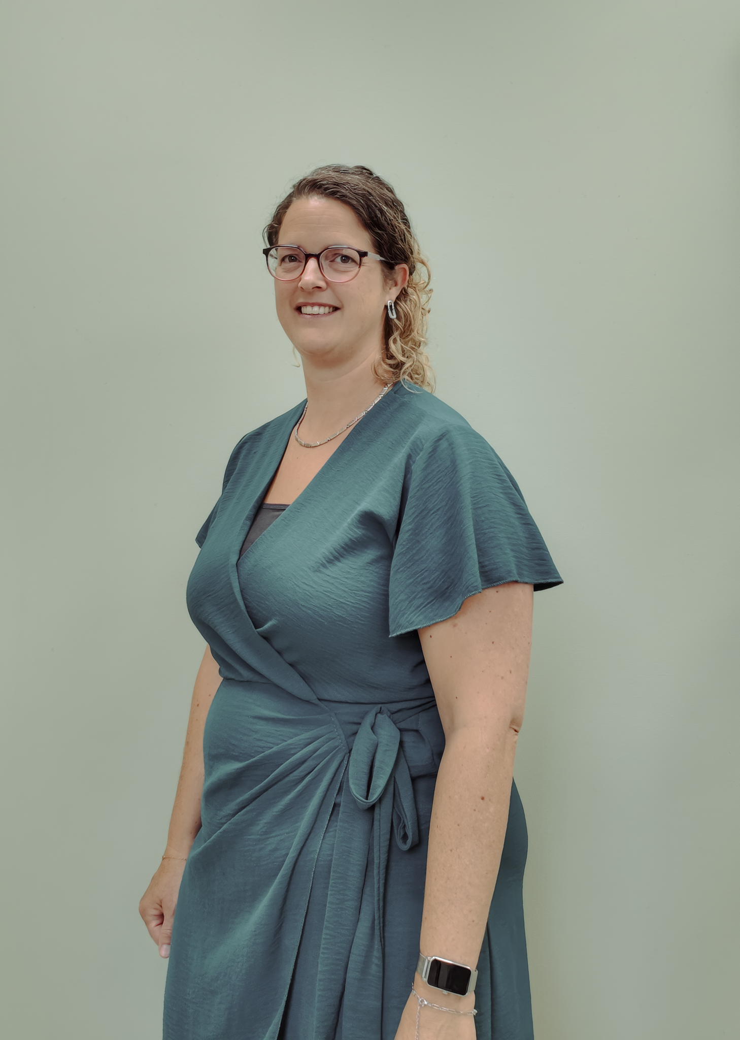 Portret van een vrouw met bruin, gekruld haar, brilmontuur en oorbellen, gekleed in een blauwe jurk, staand voor een licht groene achtergrond.
