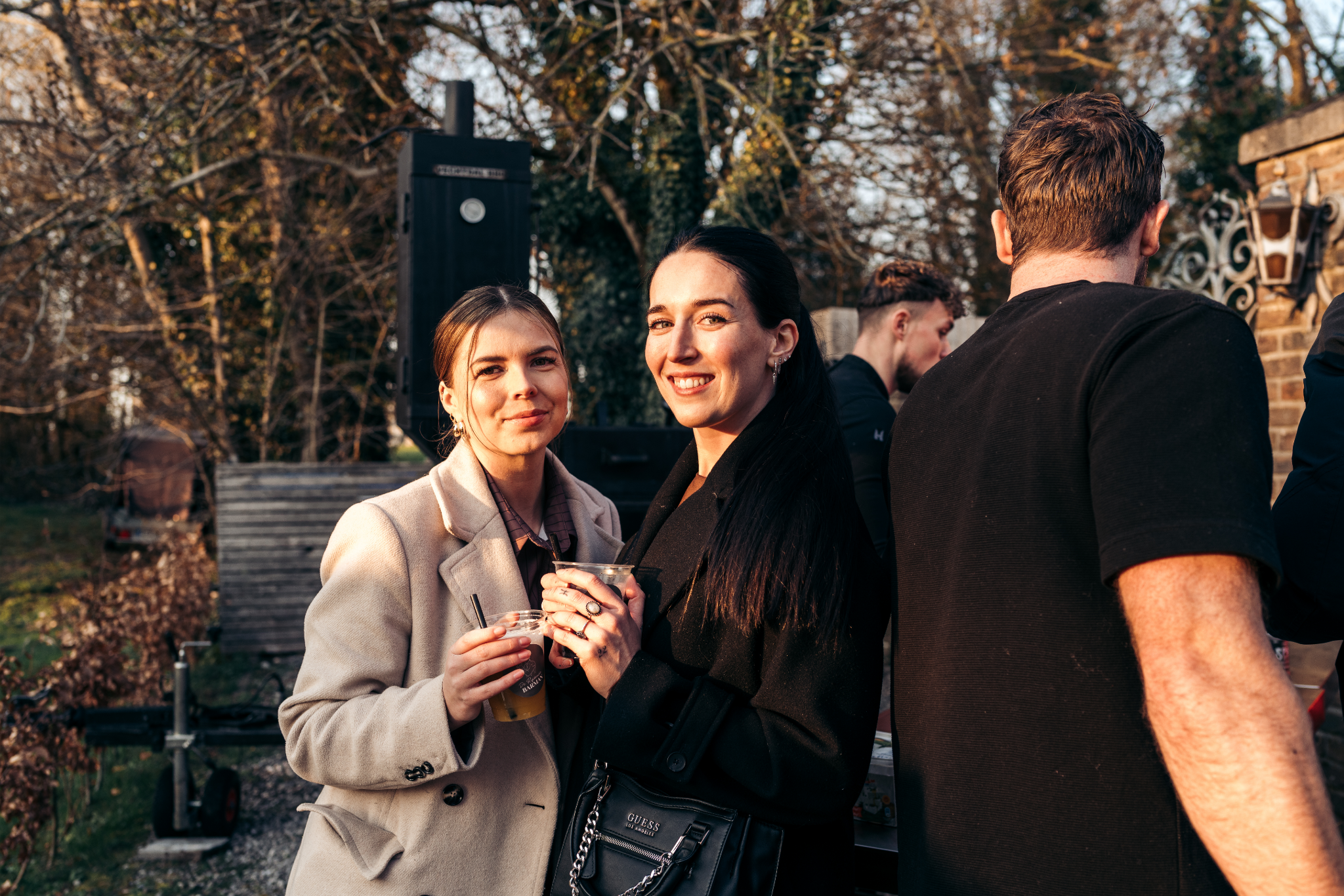 Twee vrouwen met drankjes in de hand op een buitenfeest tijdens zonsondergang, met andere mensen op de achtergrond.