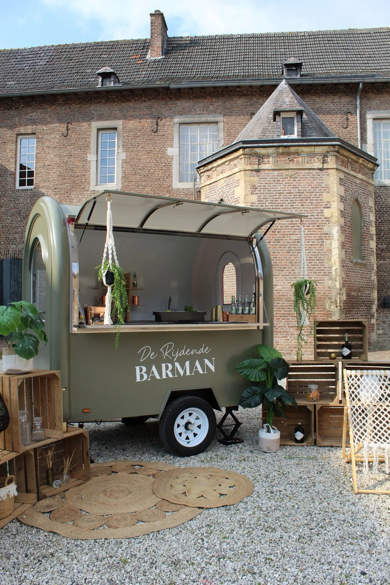 Een mobiele barwagen genaamd 'De Rijdende Barman' staat op een stuk grond met grind, omgeven door decoratieve planten, houten kratten en touwen met hangplanten, voor een oude stenen gebouw met grote ramen. Cocktails & BBQ