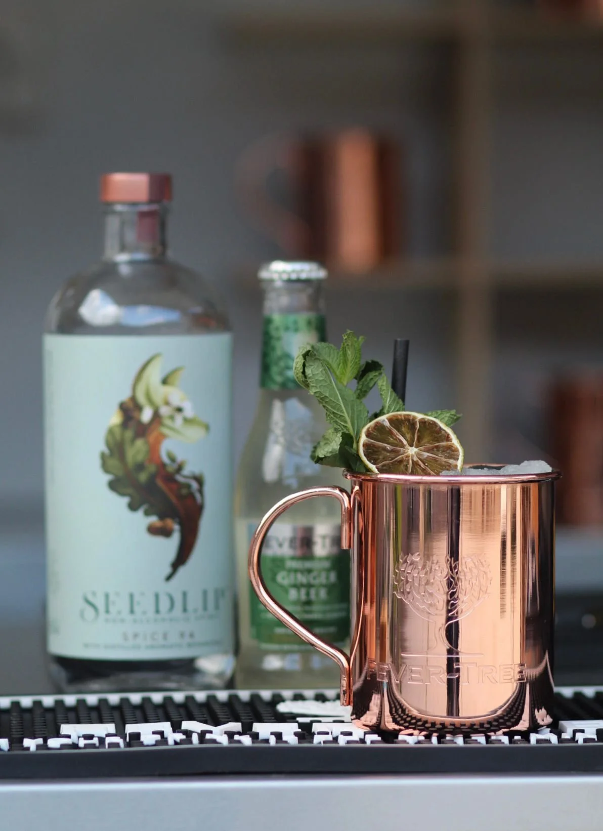 Een kop met een munt, munt, en muntplant, waarschijnlijk gember, in een kopje op een bar met drankflessen op de achtergrond.