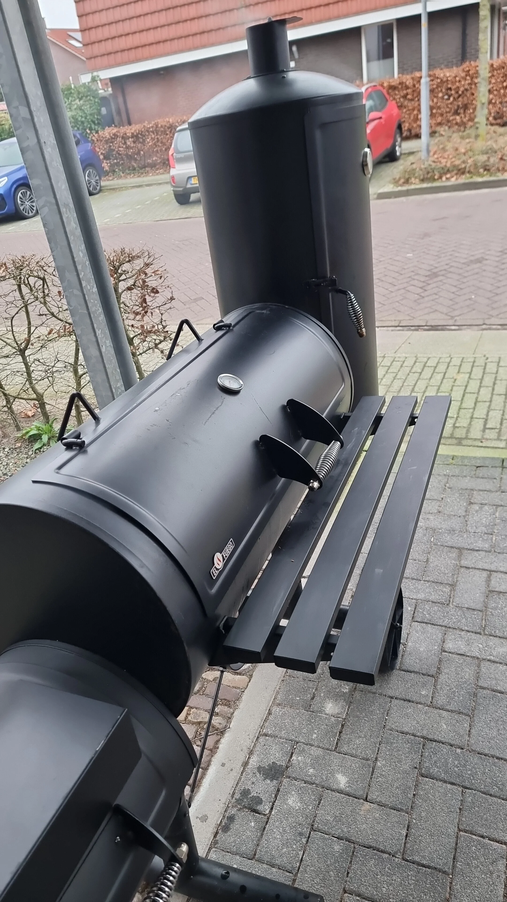 Offset barbecue smoker with chimney, parked cars in background.