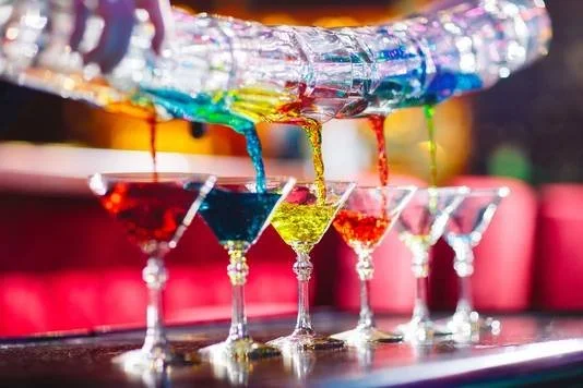 Vijf kleurrijke cocktails in martini-glazen onder een regenboog-folie met verf die druppelt.
