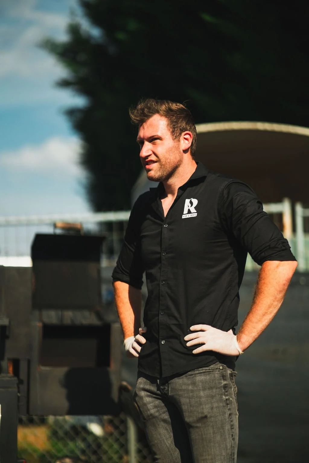 Een man met bruin haar, gekleed in een zwarte blouse met een wit logo aan de linkerborst, staat buiten tijdens daglicht met zijn handen in zijn taille, met een achtergrond van bomen en een blauwe hemel.