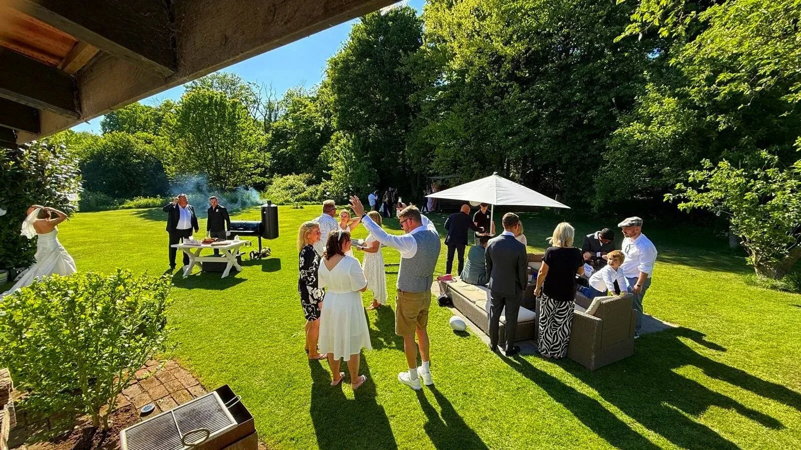 Buitendiner bij een tuin, mensen staan en zitten onder een parasol, kinderen en volwassenen genieten van een feest op een zonnige dag