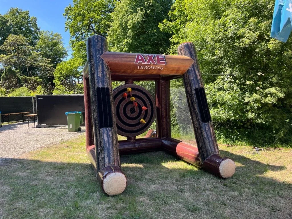 Een grote, houten speelkubus met een doelwit en pijltjes voor houten pijltjesspel op een grasveld, omgeven door bomen en een blauwe lucht.