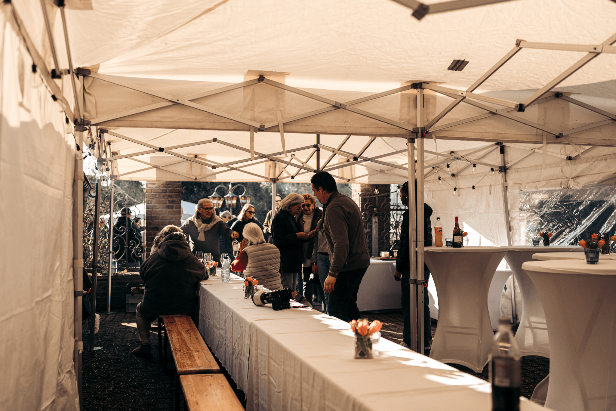 Gezellige bijeenkomst onder een witte tent met mensen die praten en drinken, tafels met bloemen en drankflessen, in een buitenruimte met stenen muur en hekwerk.