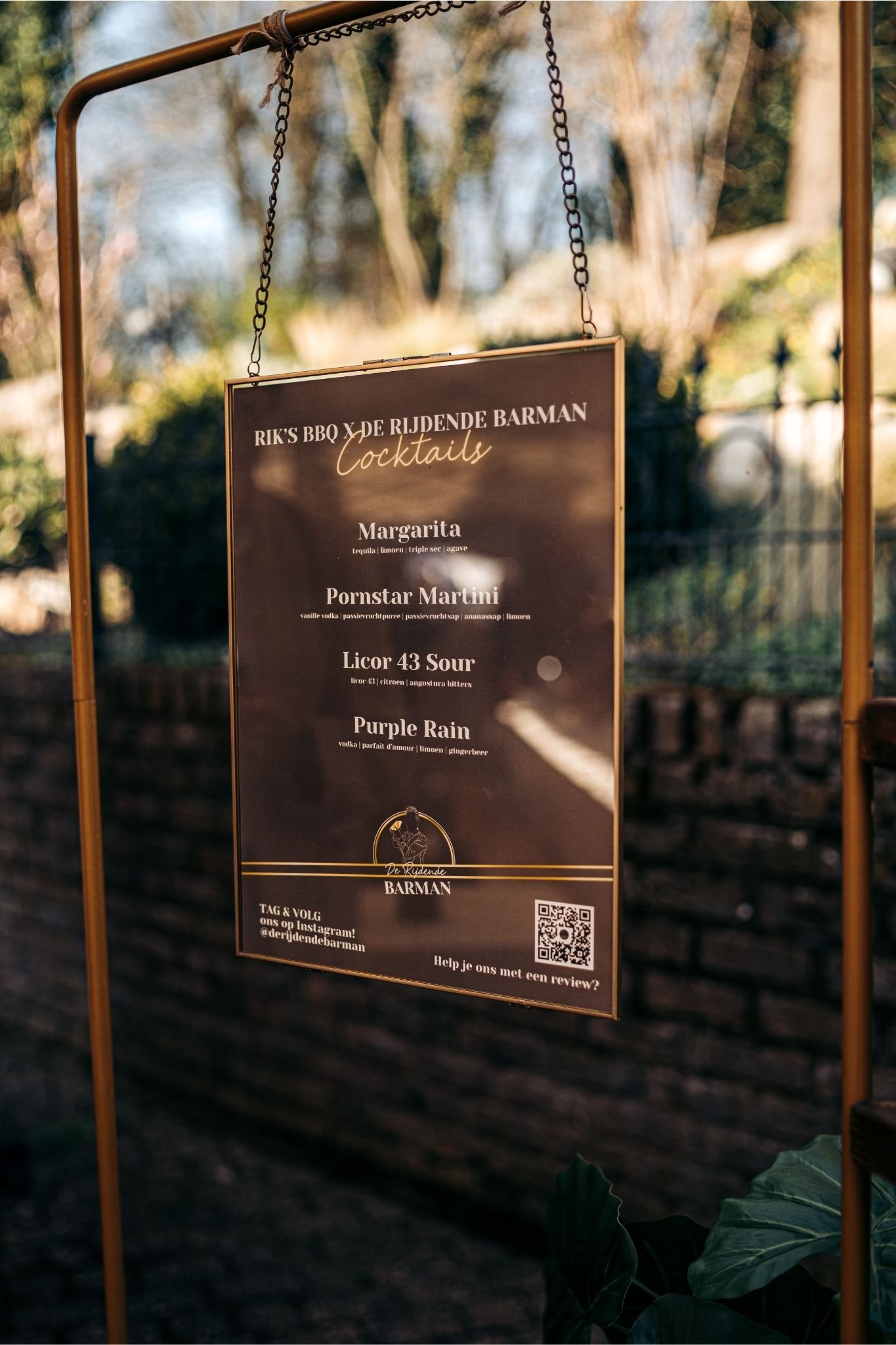 Menukaart met cocktailopties zoals Margarita, Pornstar Martini, Licor 43 Sour en Purple Rain, hangend aan een gouden frame buiten. Positieve achtergrond met bomen en een stenen muur.