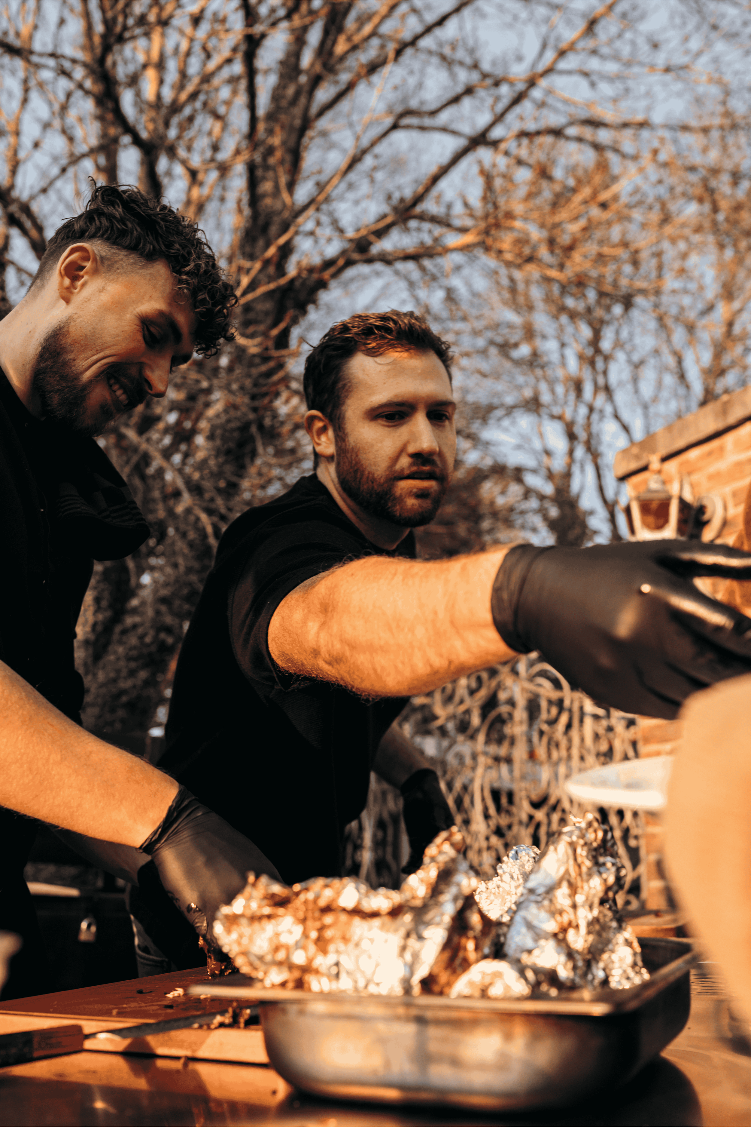 Twee mannen bereiden voedsel in een buitenomgeving, omringd door bladerige bomen, met een BBQ of grill en aluminium folie.