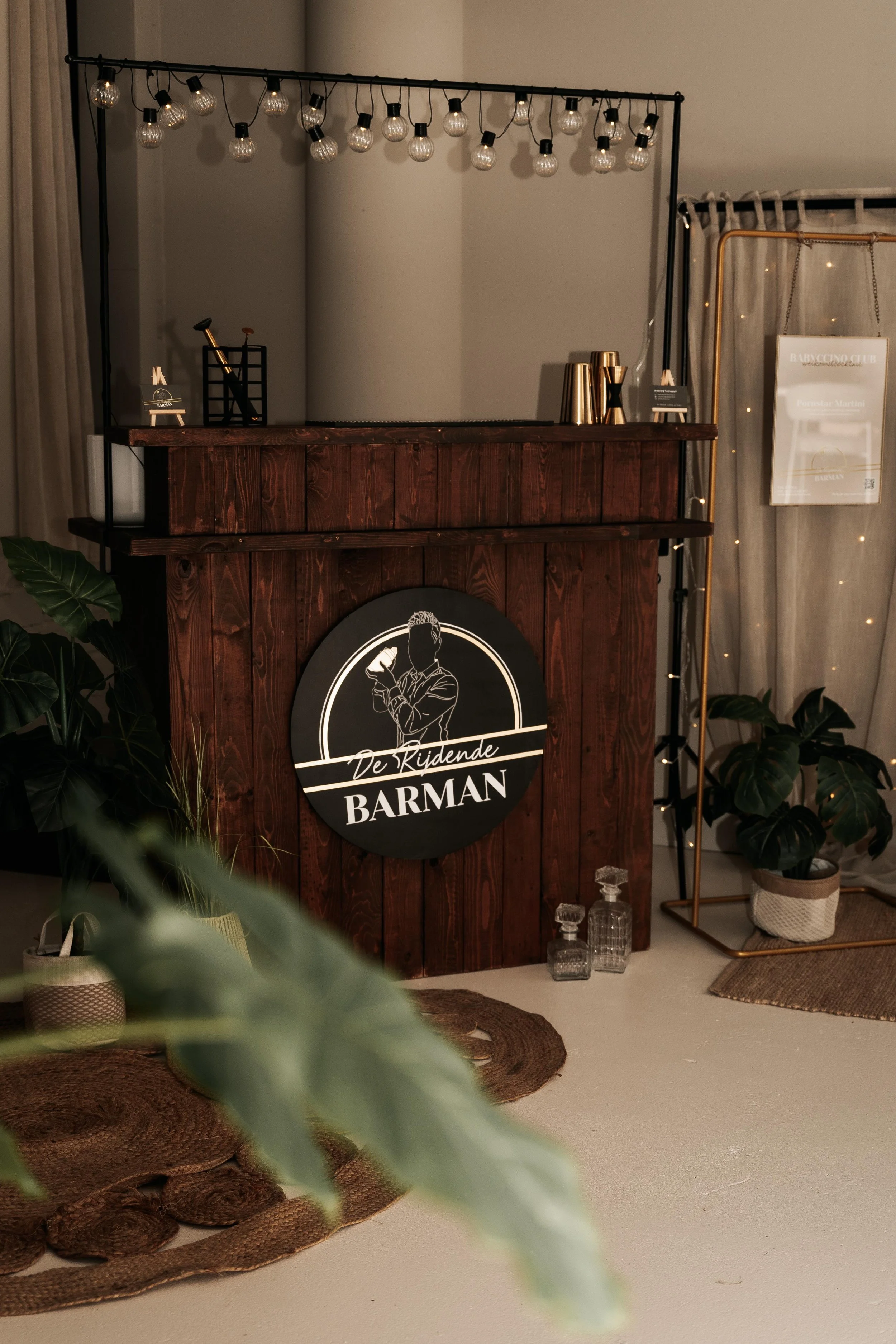 Bar met zwarte en gouden cocktailaccessoires, achtergrond met hanglampen, potted planten, en een bord met de tekst 'De Kruidende Barman'.