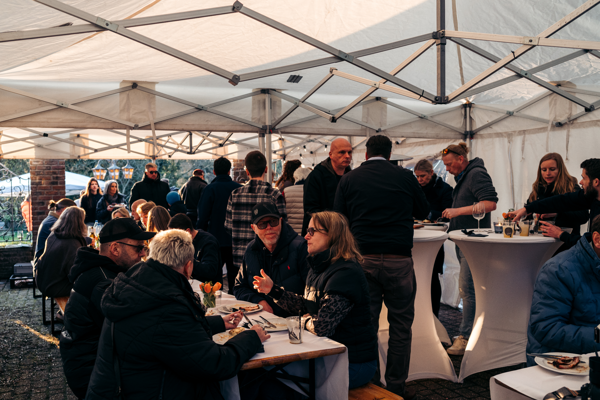 Mensen die samen eten onder witte tenten tijdens een evenement, met enkele tafels en drankjes op de voorgrond.