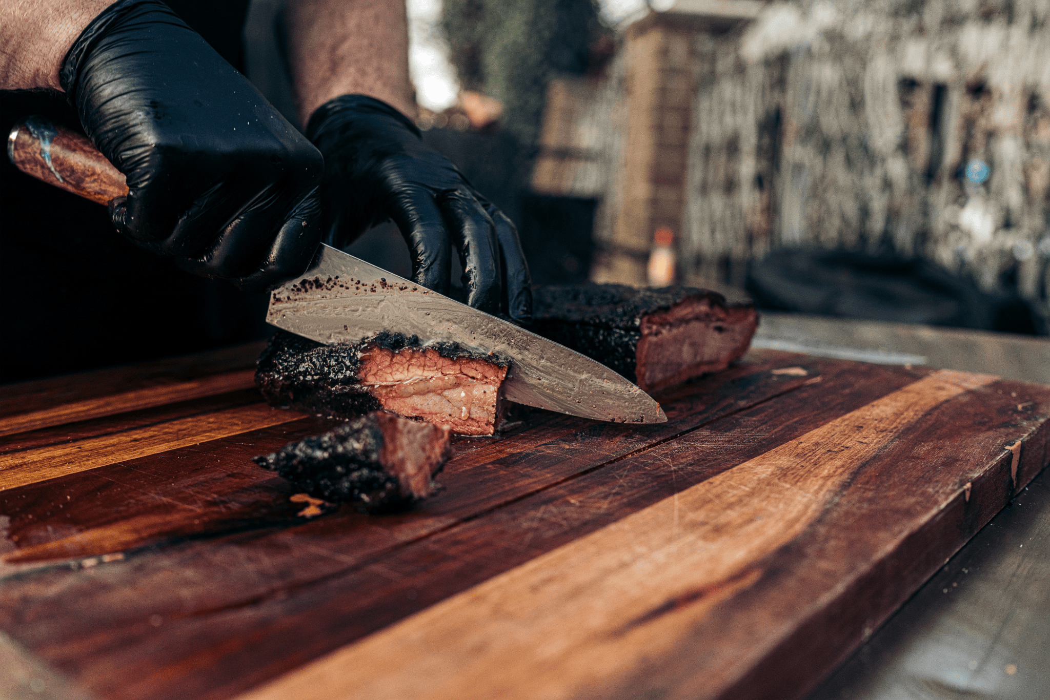 Persoon snijdt gerookt vlees op een houten snijplank, vingers bedekt met zwarte handschoenen.