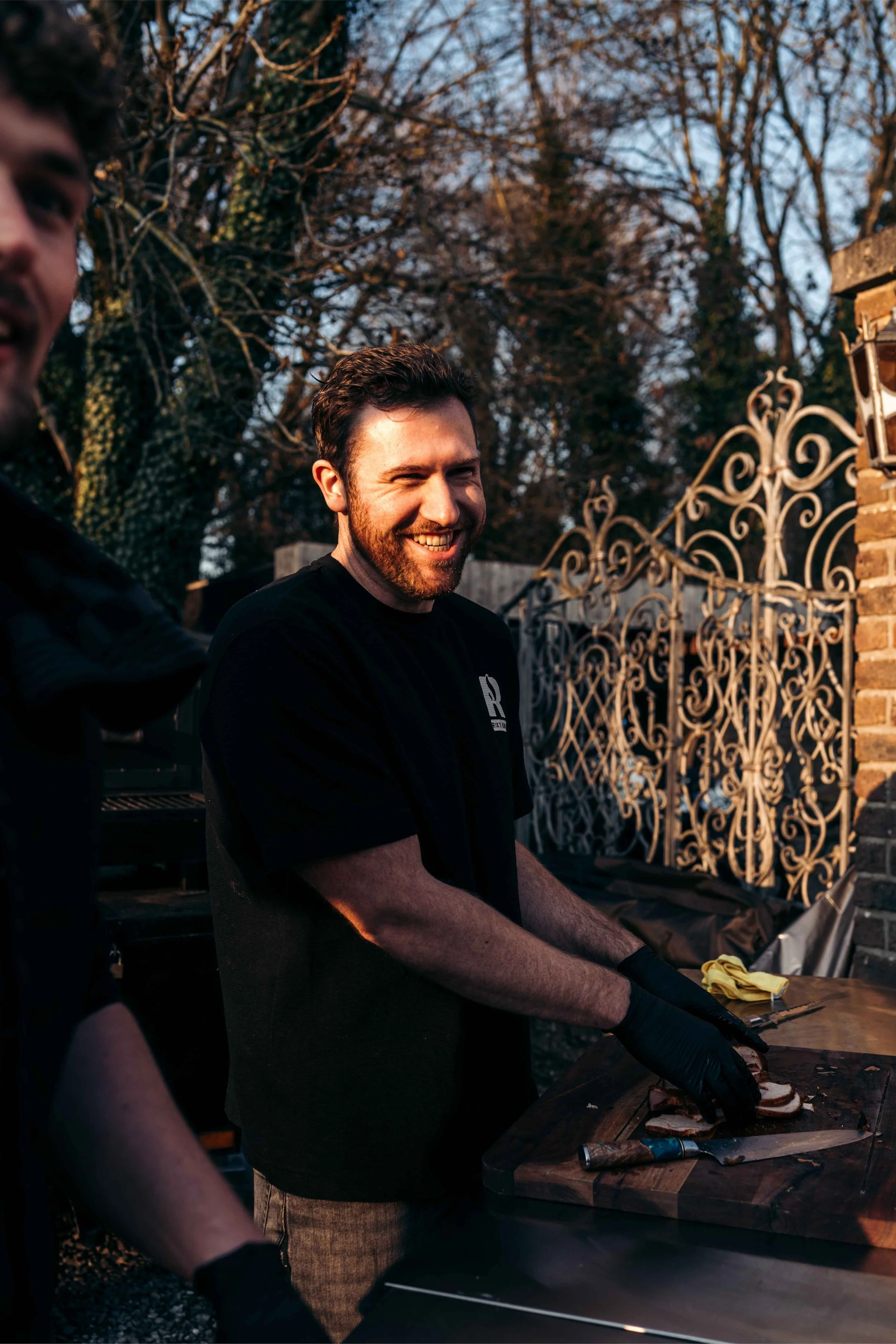 Manen bakken vlees op een houtskoolbarbecue buiten, met een background van een smeedijzeren hek en bomen, tijdens zonsondergang.