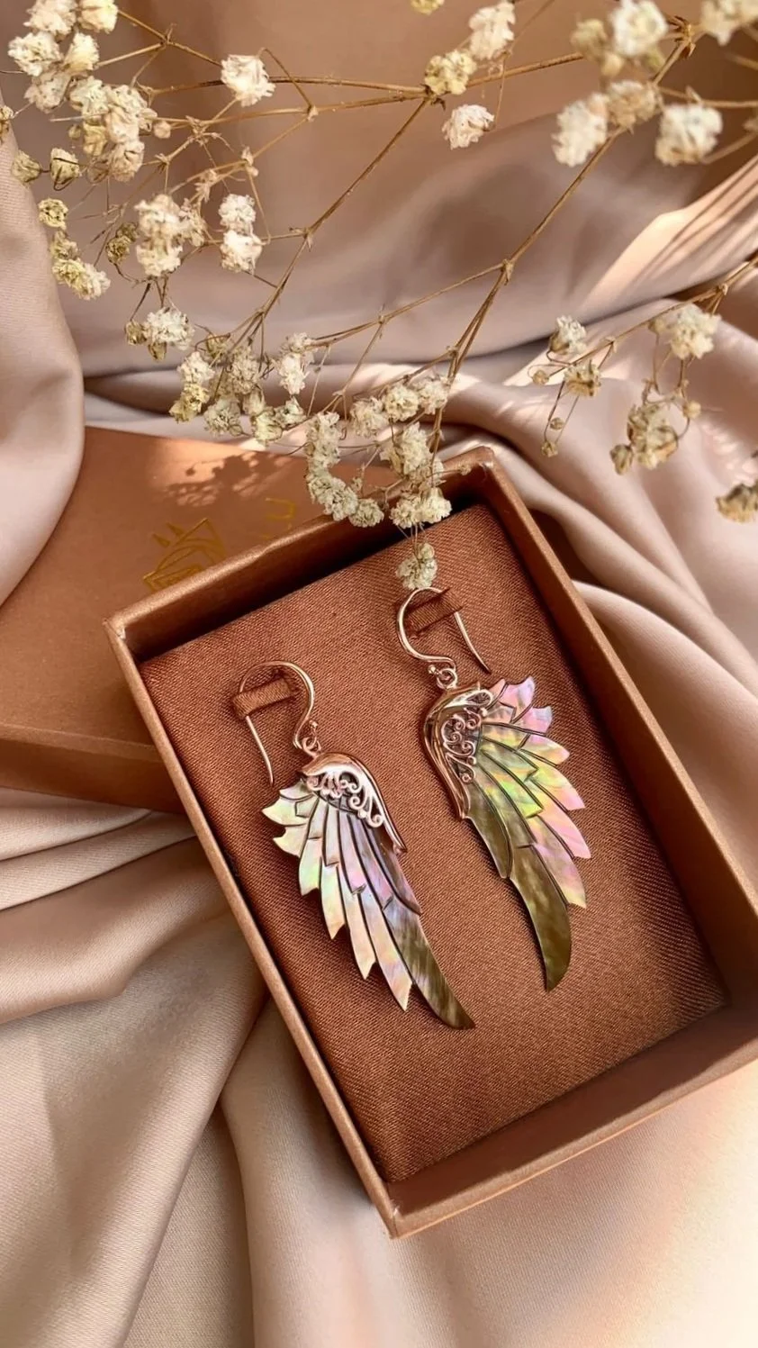 A pair of iridescent angel wing earrings inside a brown jewelry box, with white dried flowers and soft satin fabric around it.
