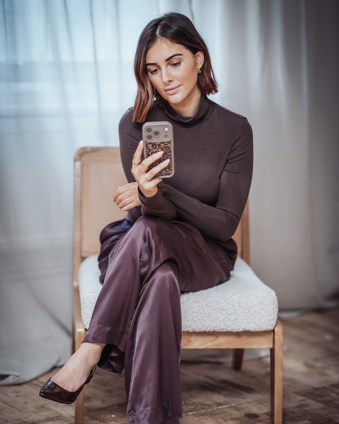 A woman with shoulder-length brown hair and earrings, dressed in a dark long-sleeve top and satin pants, is sitting on a wooden chair with a cushioned seat, looking at her phone with a leopard print case.
