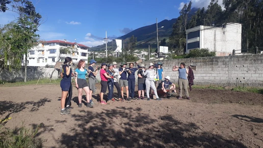 Group of people outdoors on a dirt lot with mountains and buildings in the background.