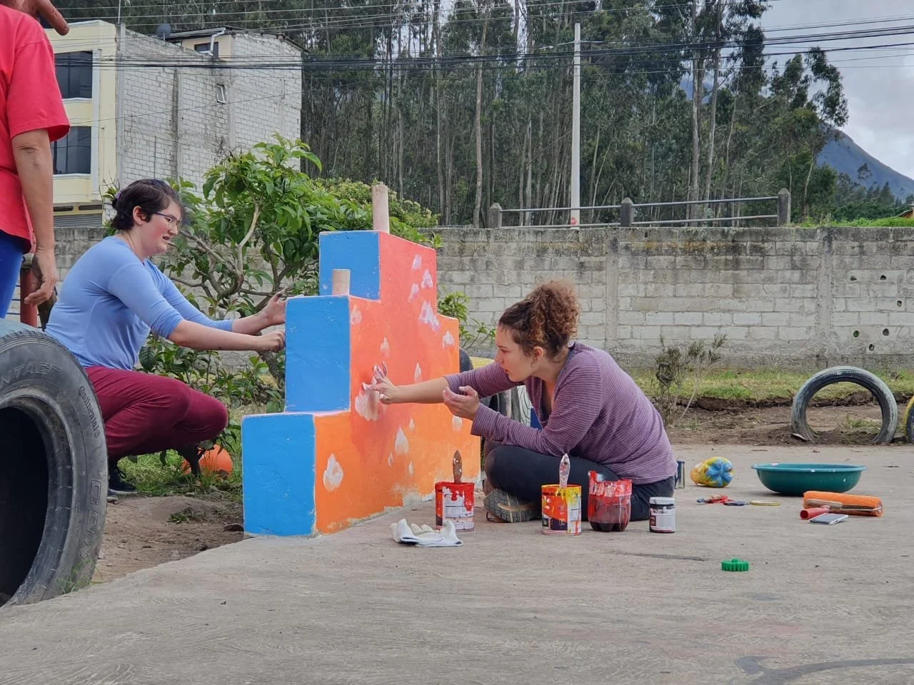 Two women are painting a colorful mural on a wall outdoors, with one sitting on the ground and the other kneeling, surrounded by paint supplies and tools.