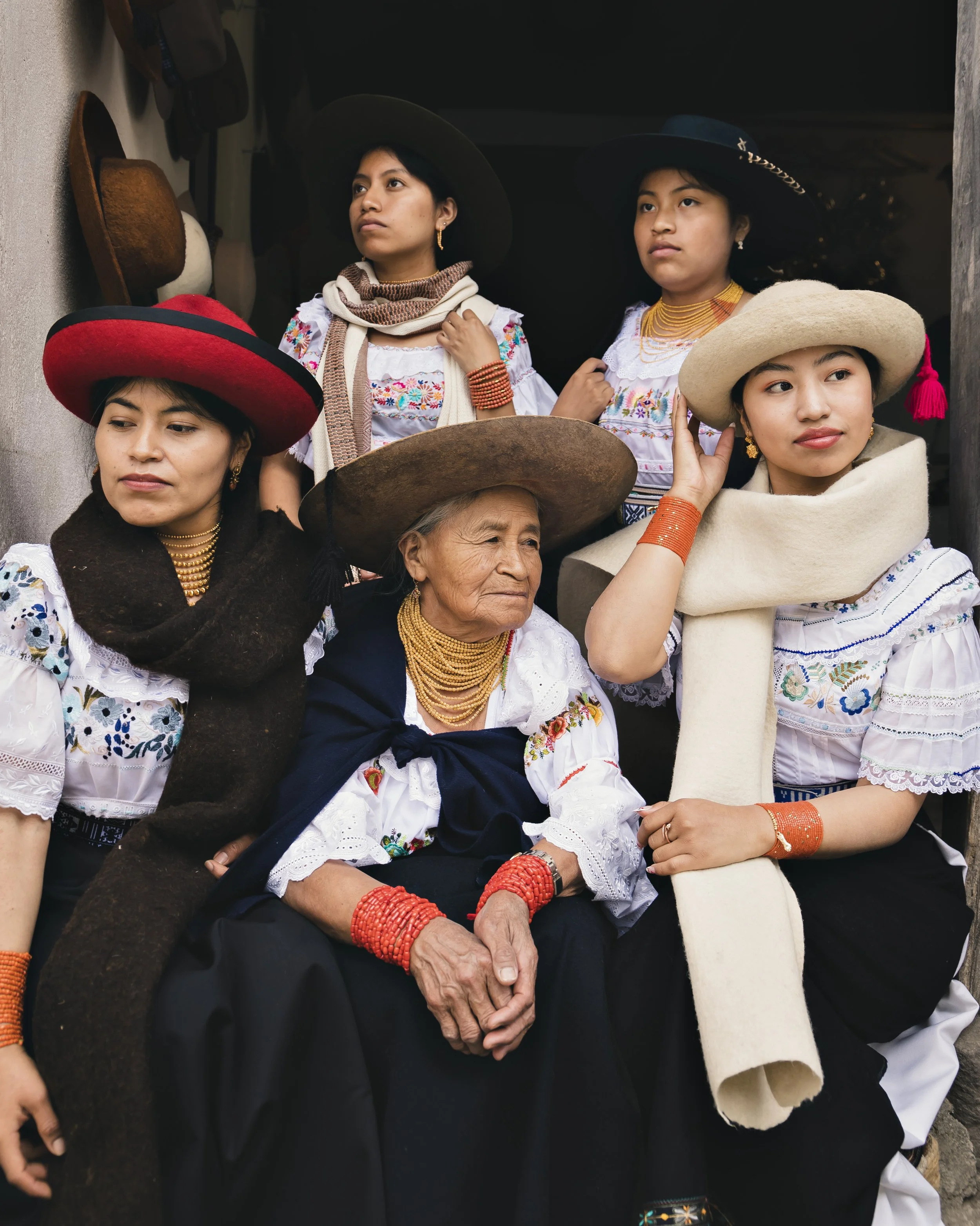 Group of women and an elderly woman dressed in traditional Ecuador Kichwa clothing, wearing embroidered blouses, colorful jewelry, and wide-brimmed hats, gathered together in a festive setting.