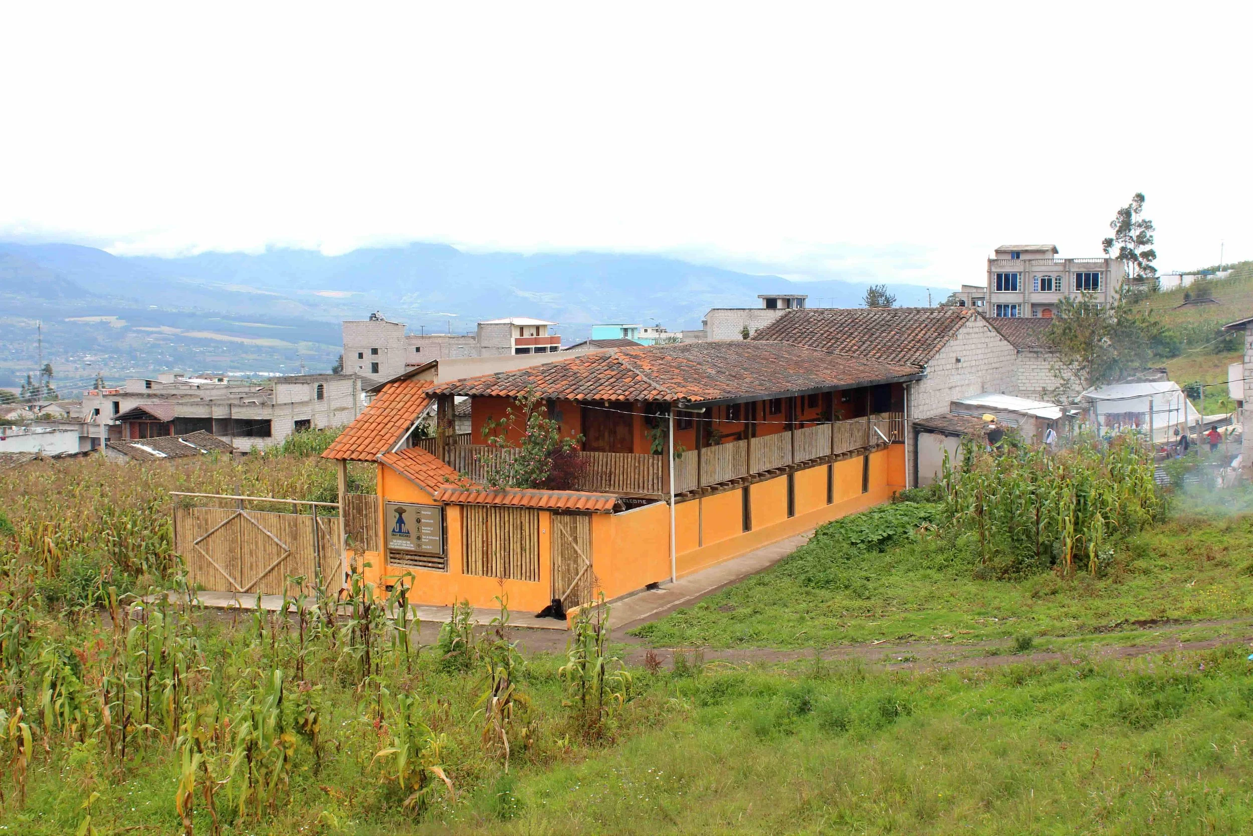 A rural house with an orange exterior and a tiled roof, surrounded by a garden and a field of corn in a hilly area with a small town nearby and mountains in the background.