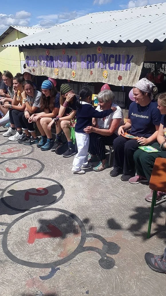 Children and adults sitting outdoors on a curb in front of a building with a banner. A young girl is embracing an older woman. The ground has painted numbered circles, and there are balloons and a small table nearby.
