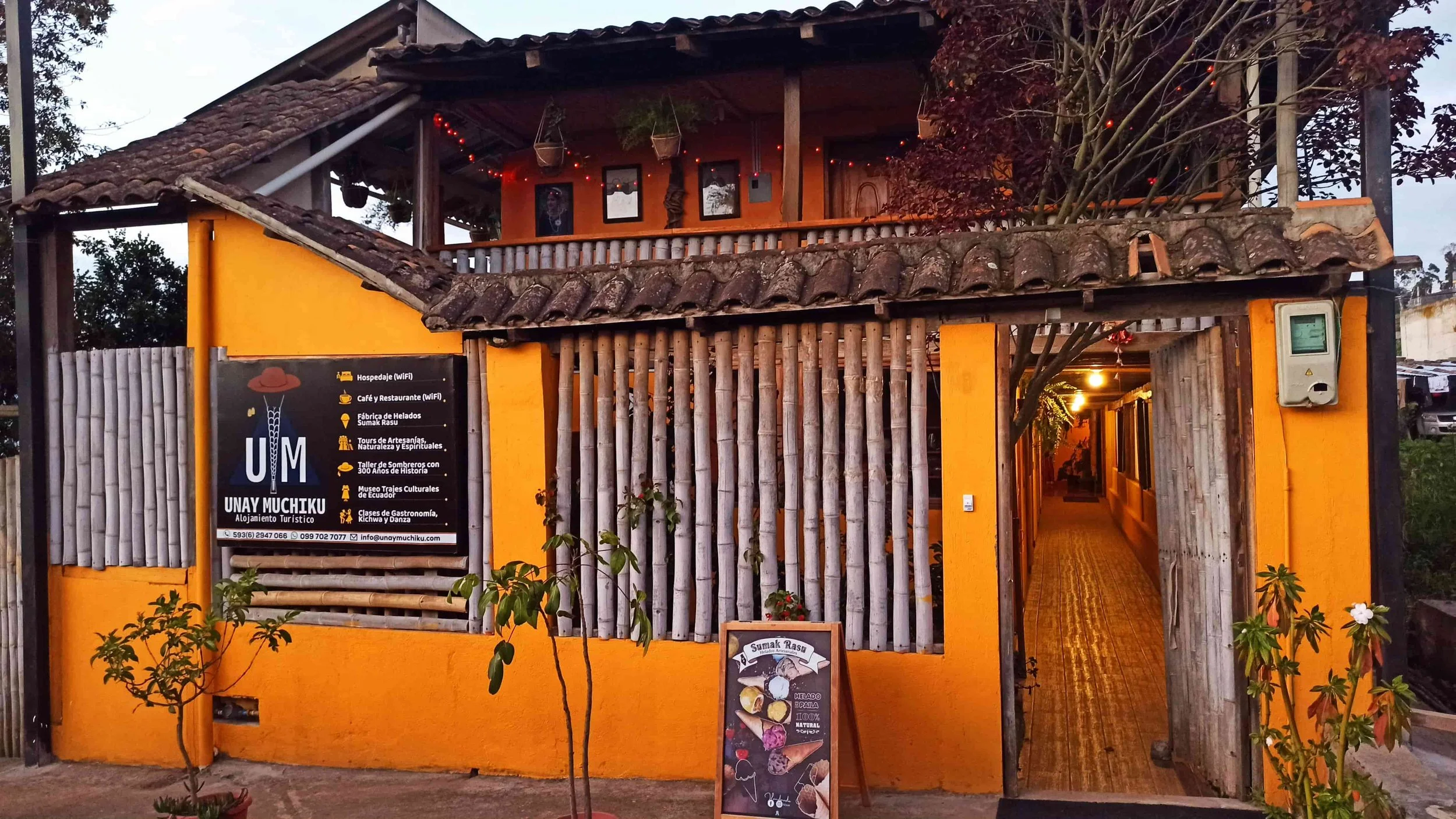 A two-story building painted orange with a wooden balcony on the upper floor. The entrance features a wooden archway, and there are plants and a signboard outside. The signboard advertises accommodations, coffee, an ice cream shop, and cultural tours in Ecuador.