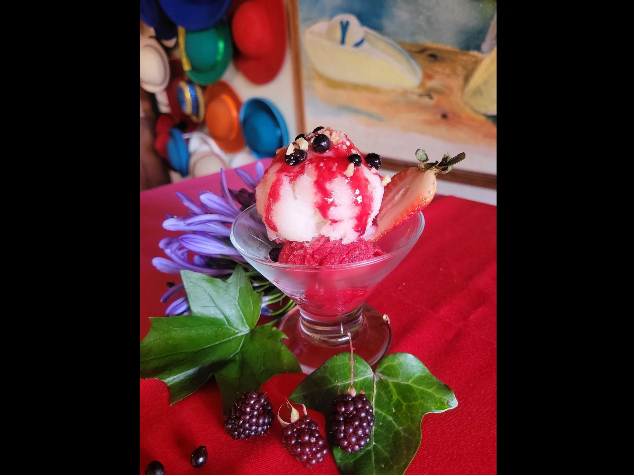 A glass bowl of fruit sorbet with blackberries and a strawberry garnish, surrounded by green leaves and a purple flower on a red cloth, with a colorful background of containers and a painting.