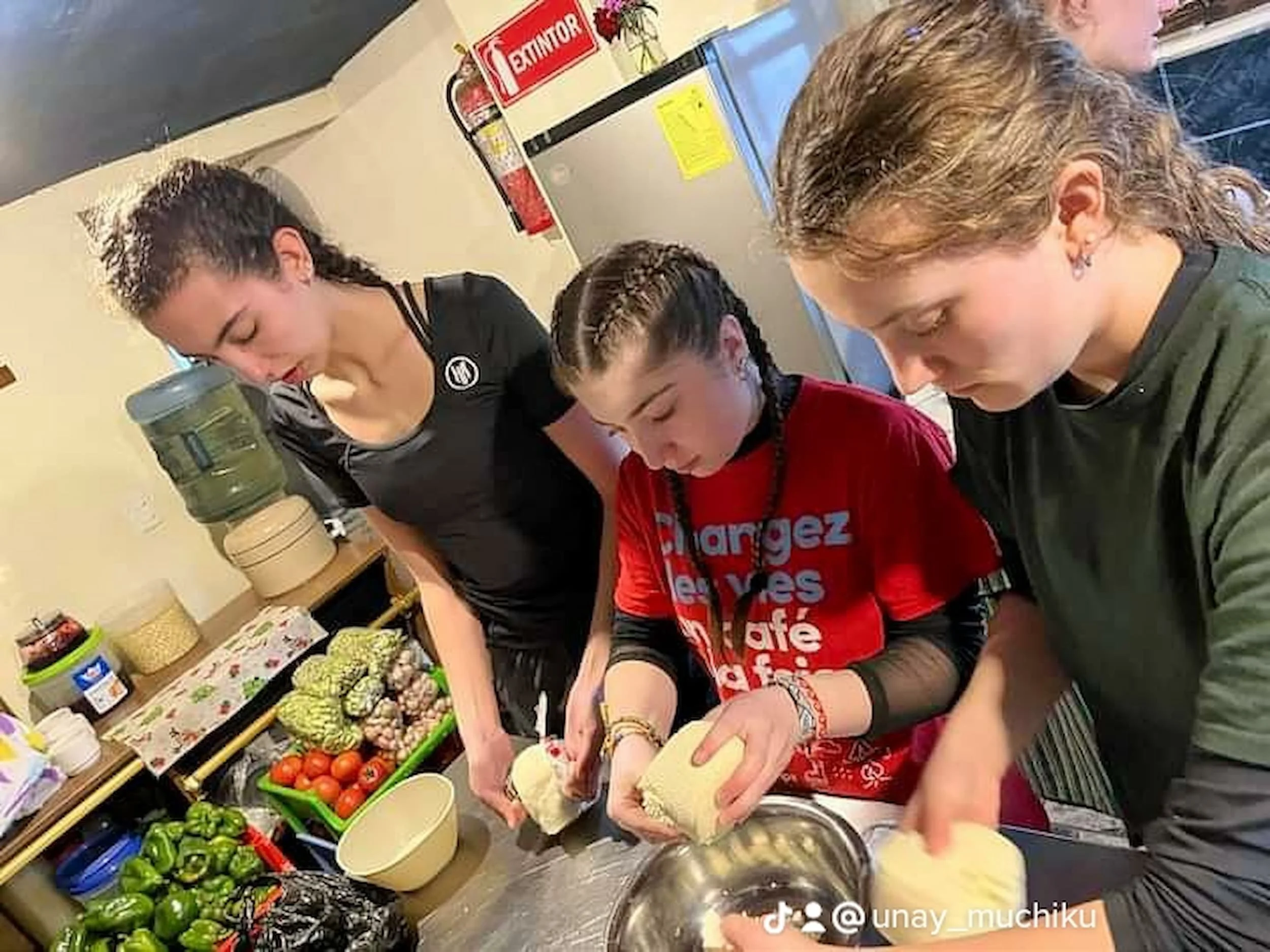 Three young girls are preparing food together in a kitchen, peeling or cutting vegetables at a table filled with produce, including tomatoes and green peppers.