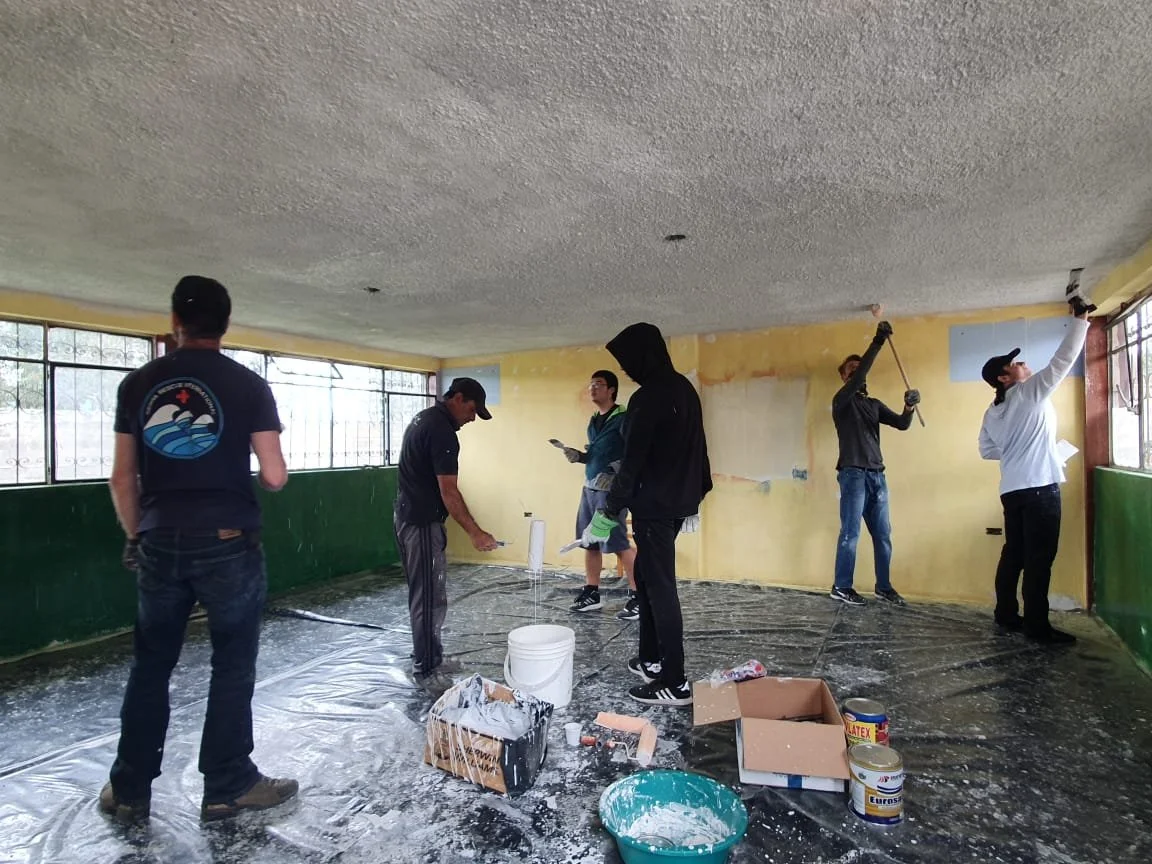 Group of people renovating a room, painting and repairing the walls, with painting supplies and plastic covering the floor.