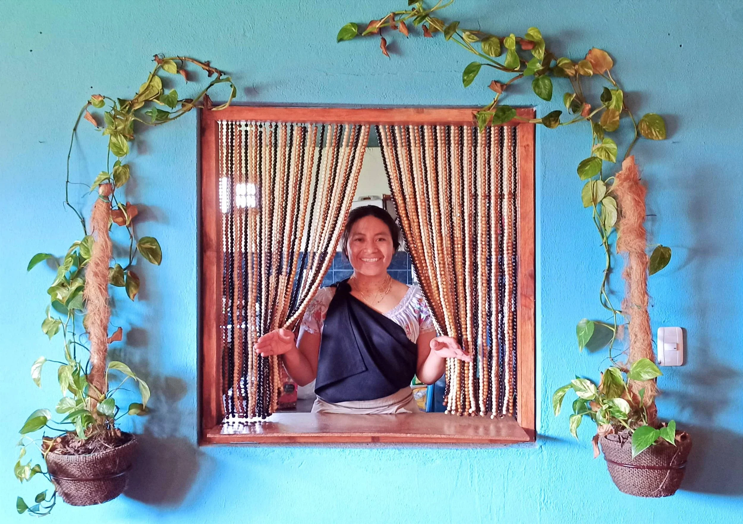 A woman smiling and holding beaded curtains in a window with beaded curtain, surrounded by potted vines on a teal wall.