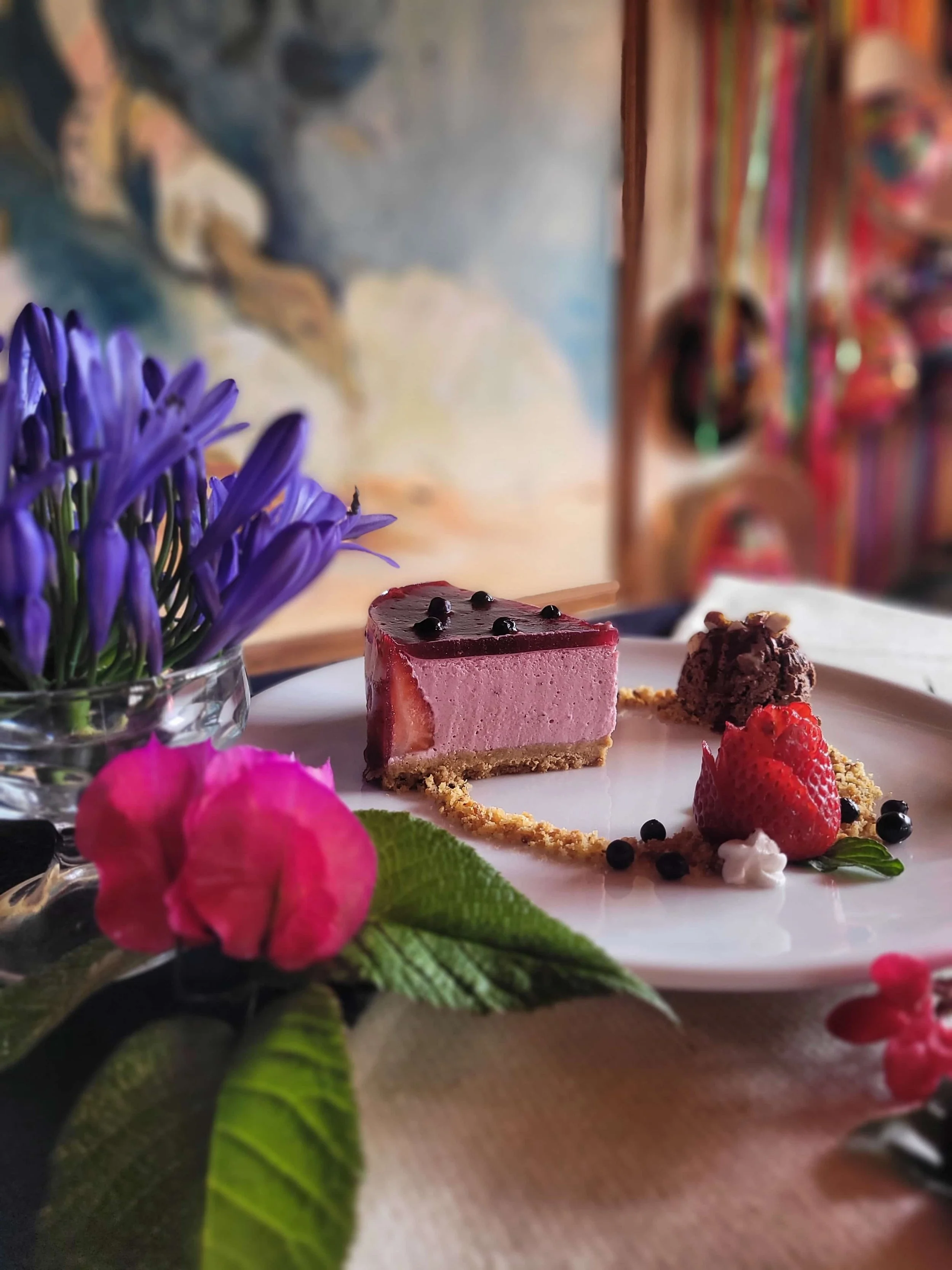 A small slice of pink cheesecake with a berry topping served on a white plate, garnished with strawberries, a chocolate bite, strawberries, blueberries, a dollop of whipped cream, and a green leaf, with purple flowers in a vase nearby.