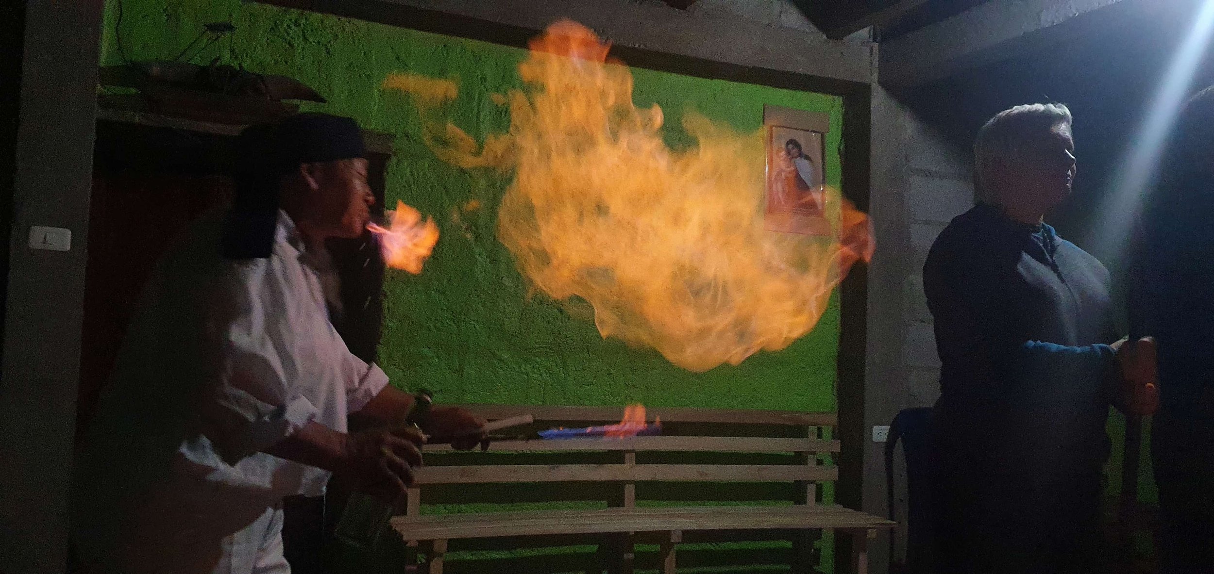 A person in white clothes and a black head covering is creating fire with a torch, producing a large flame. Another person with short gray hair and dark clothing stands nearby in a room with a bright light source, green and gray walls, and a bench.