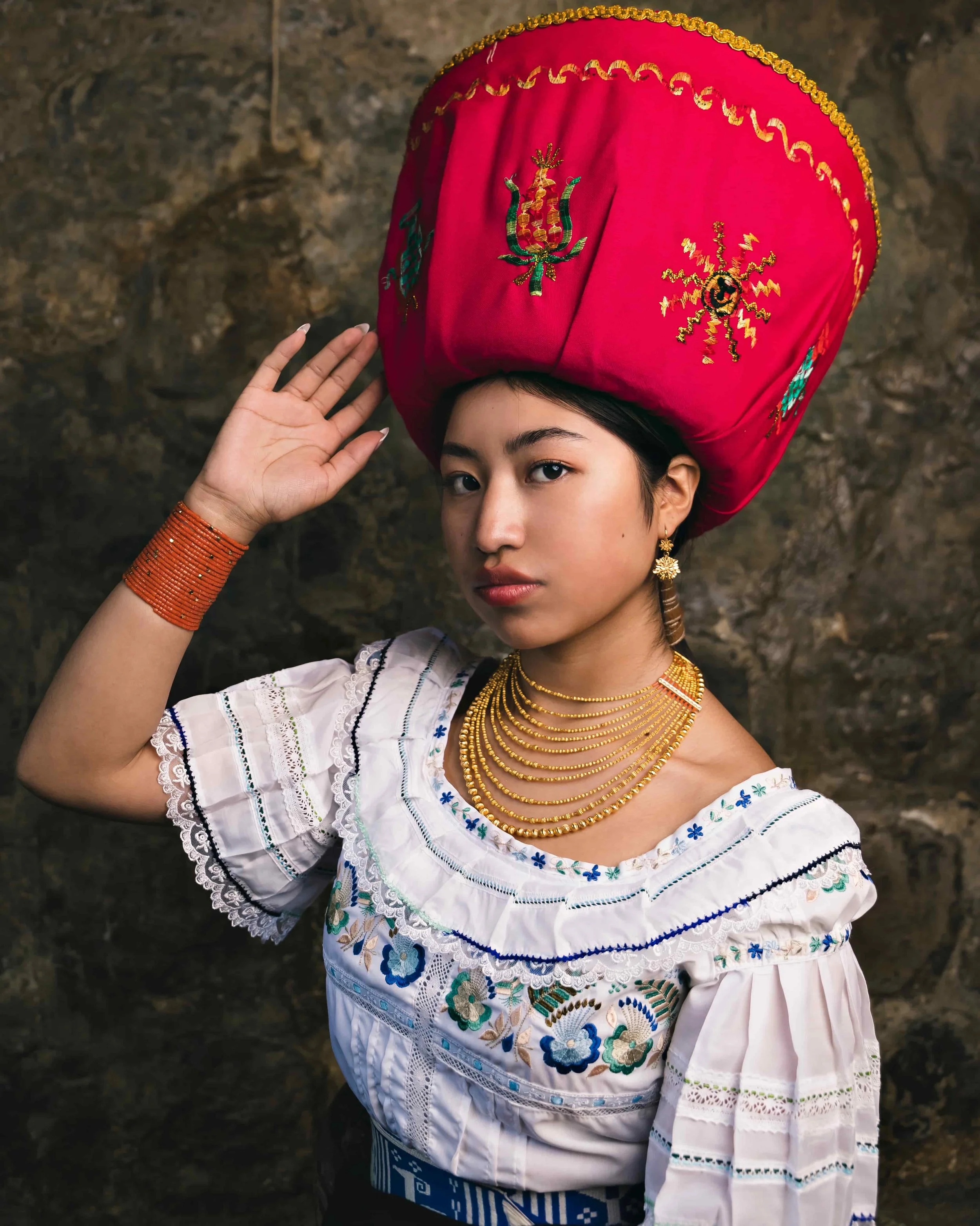 A woman wearing traditional Ecuador Kichwa attire, including a large red embroidered sombrero and a white blouse with floral embroidery, with gold jewelry and a brown earring, posing against a rocky background.