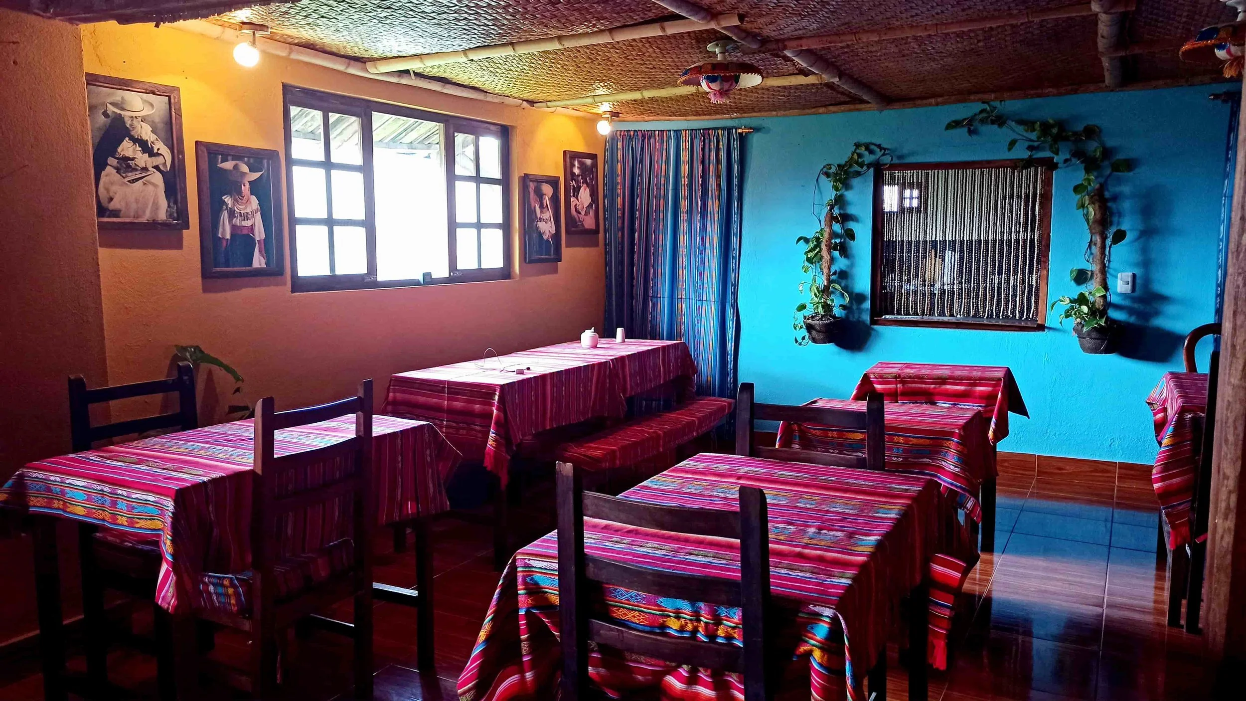 Colorful restaurant interior with tables covered in red striped tablecloths, wooden chairs, blue and yellow walls, framed pictures of people in traditional Ecuador attire, and potted plants.