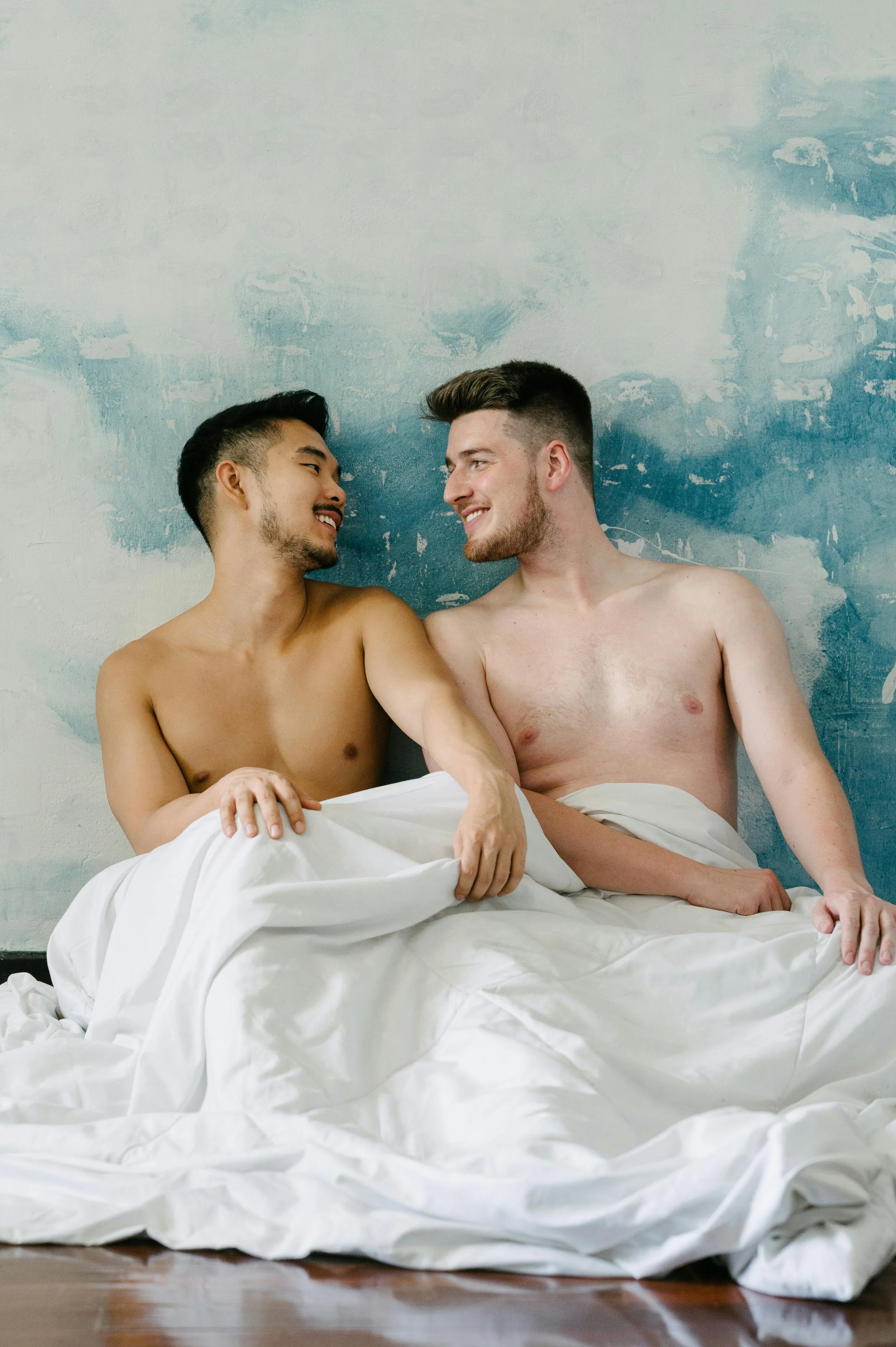 Two adult men sitting closely together under a blanket, sharing a quiet and intimate moment