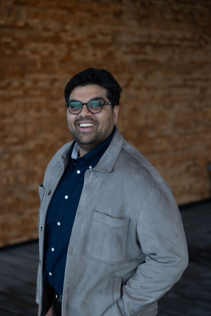 A smiling man with black hair, glasses, wearing a dark blue shirt and a gray jacket, stands in front of a brick wall.