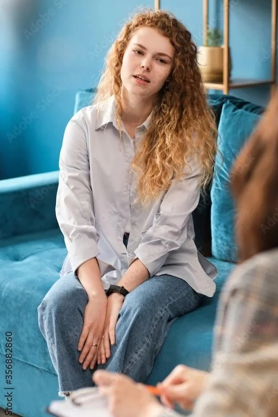 A young woman with curly red hair wearing a white shirt and blue jeans sitting on a teal sofa, appearing to be in a conversation with another person who is partially visible and holding a notepad.