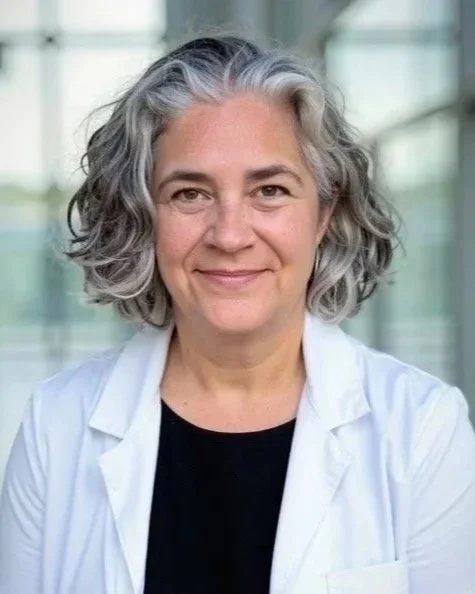 A woman with gray, curly hair smiling, wearing a white lab coat, standing in a bright, modern building with large windows.