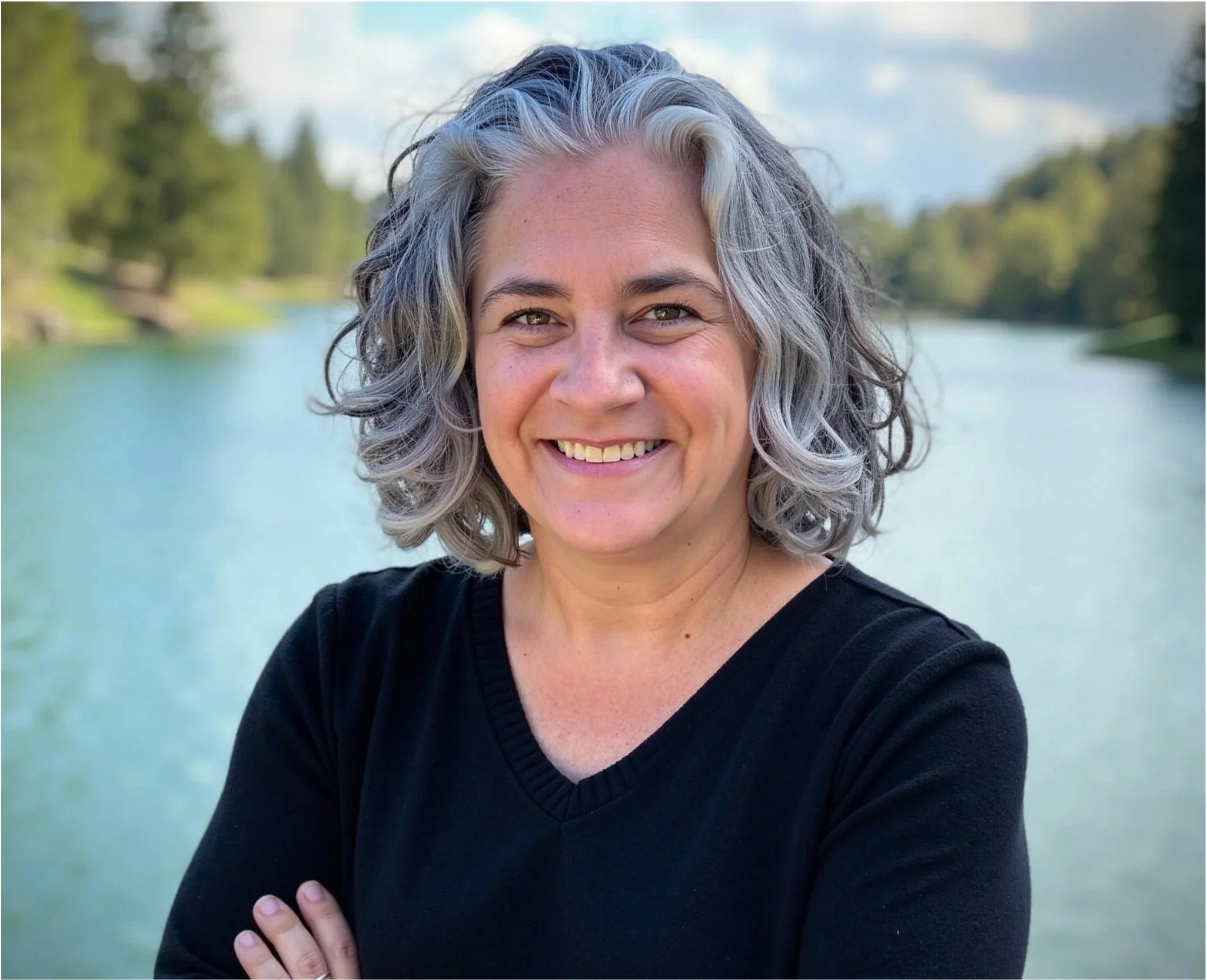 A woman with gray curly hair smiling outdoors near a lake and trees, wearing a black top.