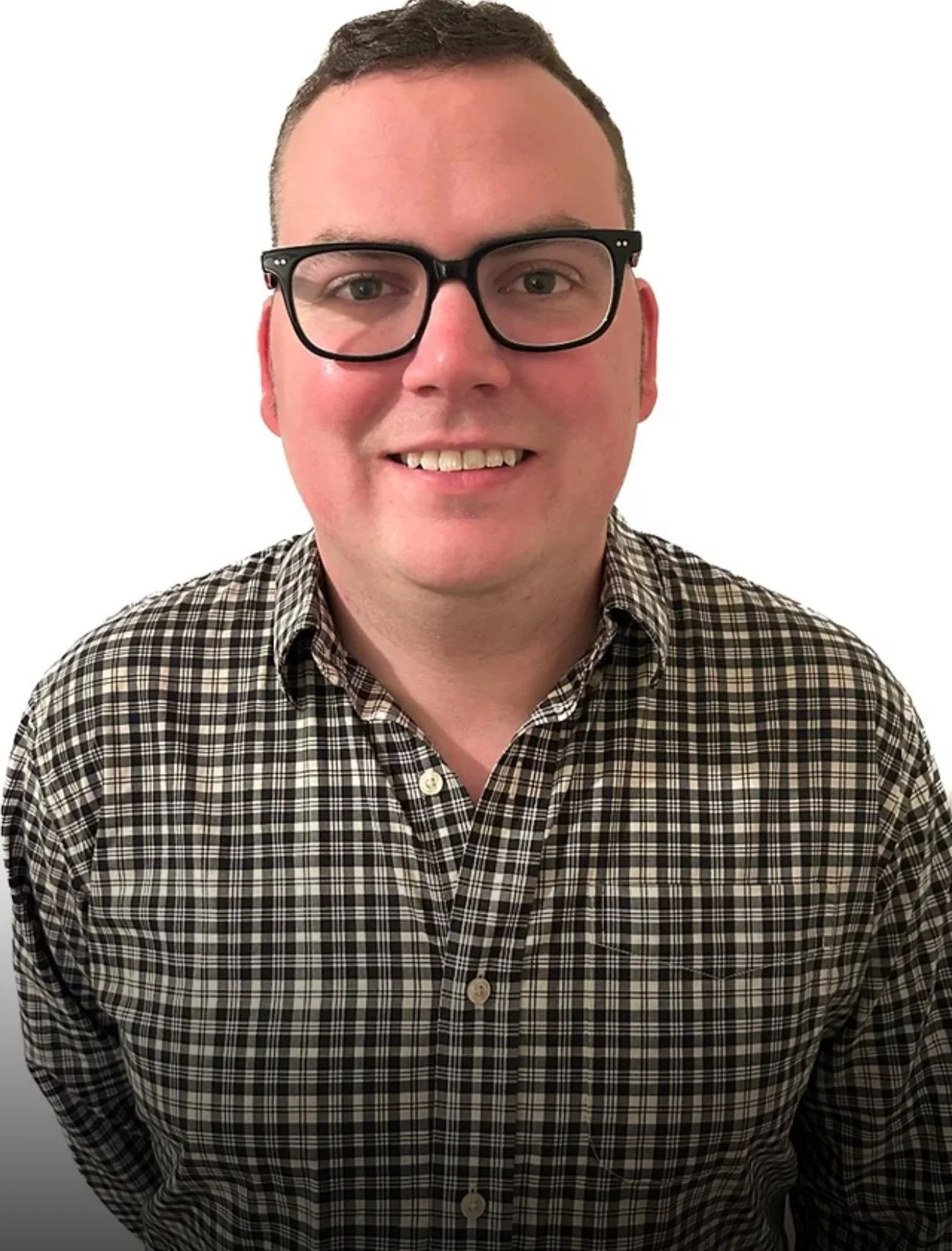 A man wearing glasses and a checkered shirt smiling in front of a plain white background.