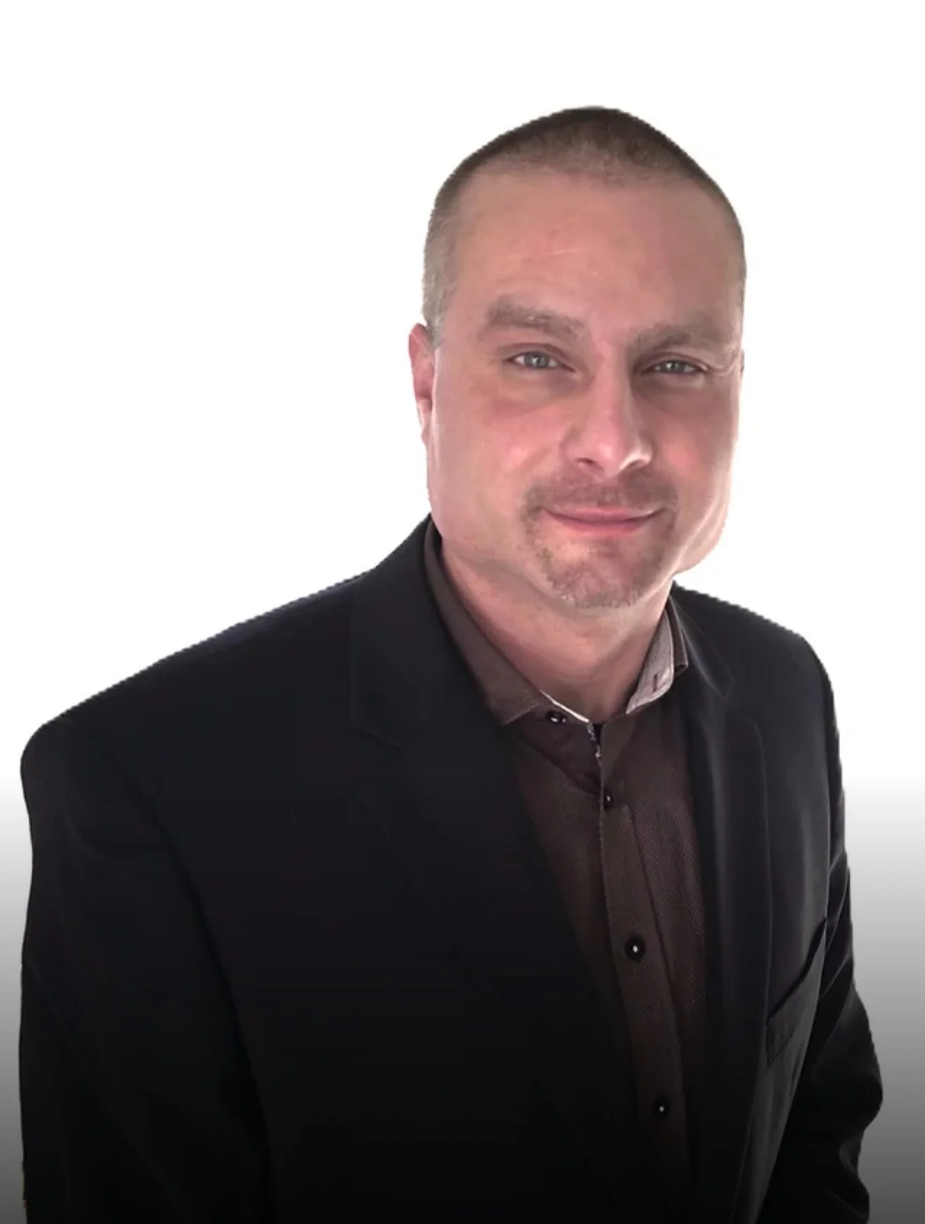 A man with a short haircut and slight beard wearing a black blazer and brown shirt, posing against a plain white background.