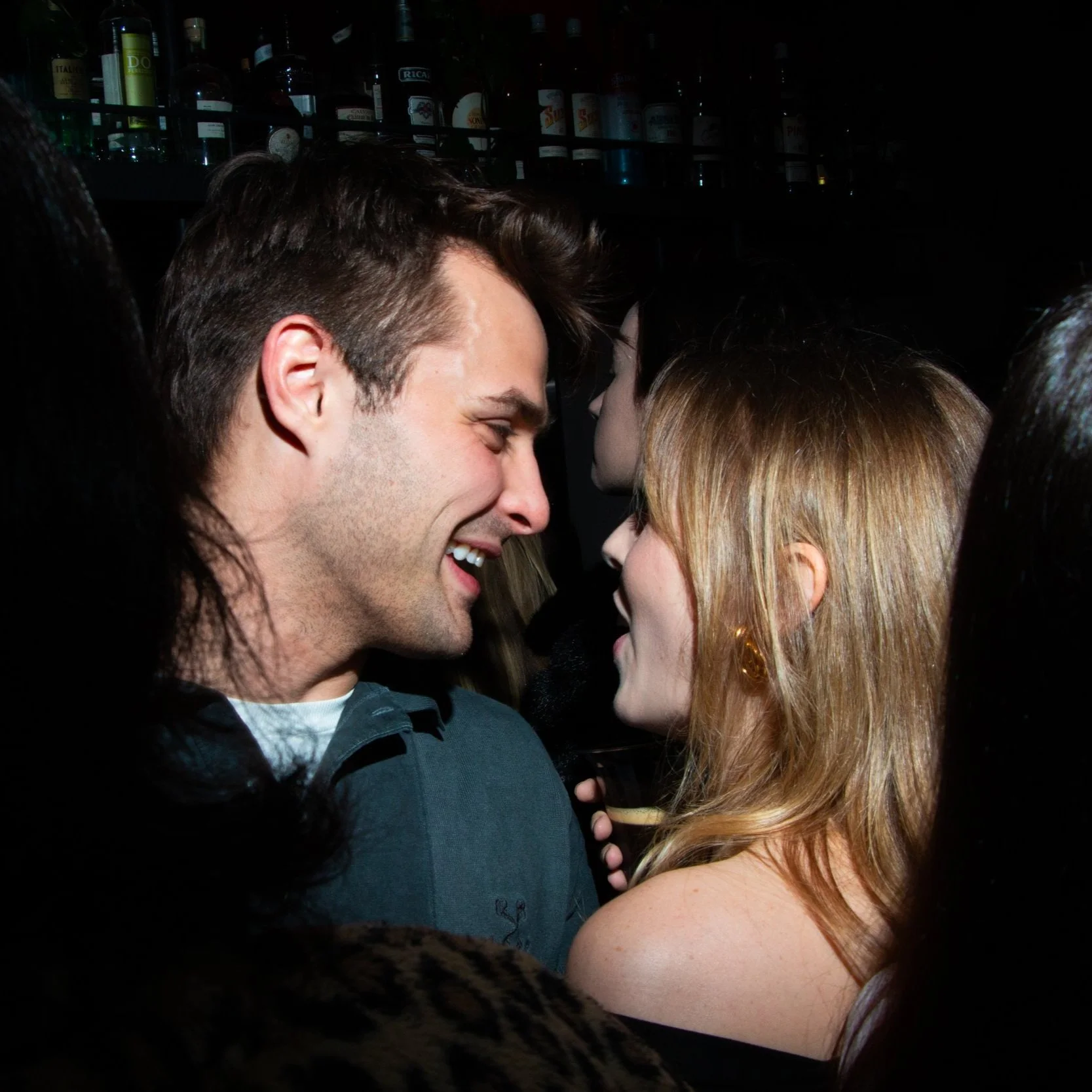 A man and woman smiling and talking closely at a crowded party with bottles visible in the background.