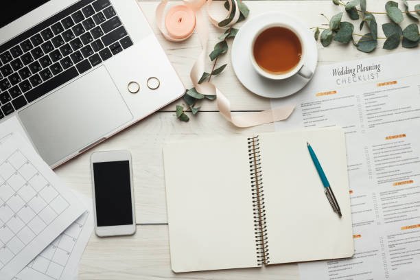 Desk setup with laptop, smartphone, wedding planning checklist, empty notebook, pen, cup of tea or coffee, eucalyptus leaves, and decorative ribbon on a white surface.