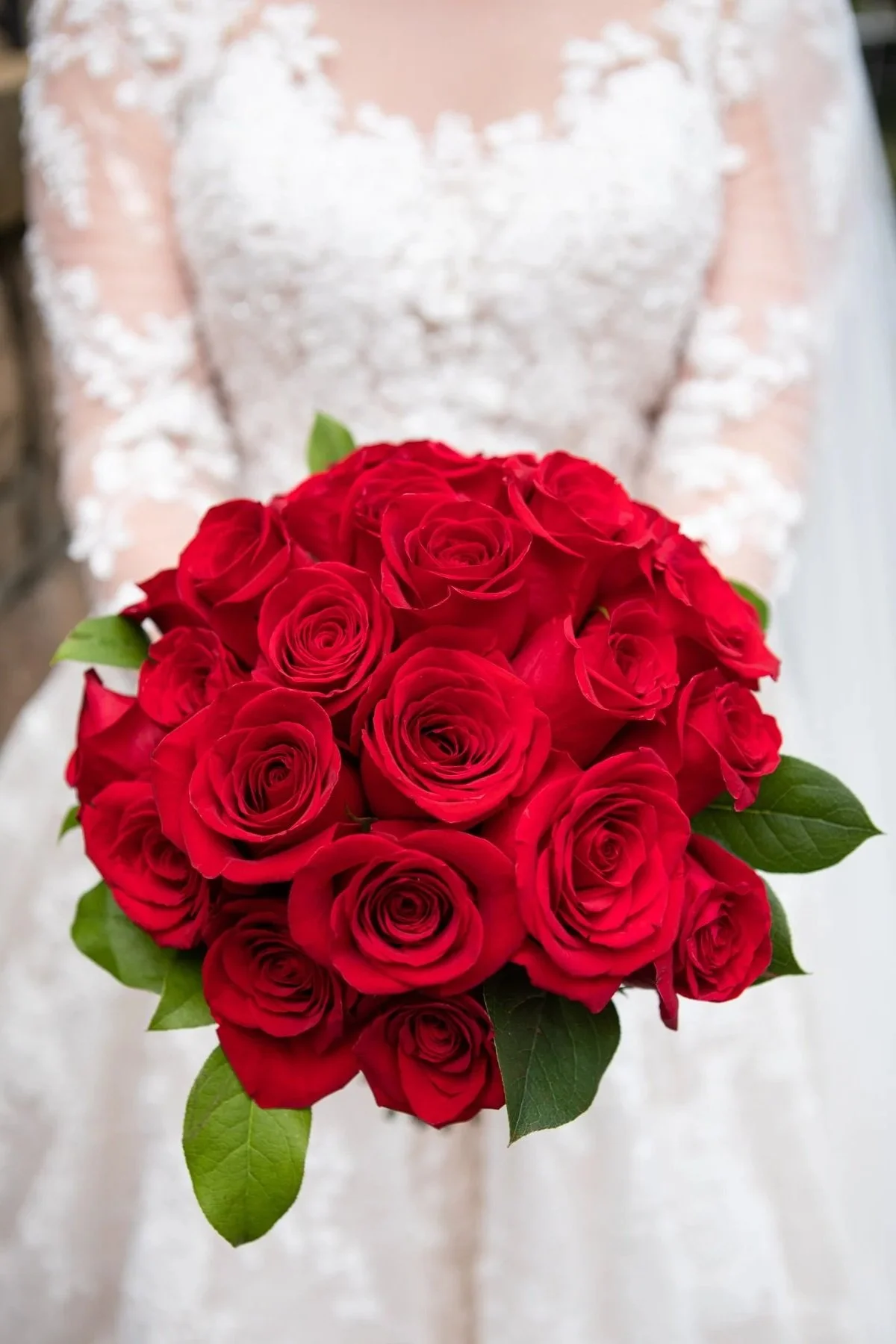 Bride in white lace dress holding a bouquet of red roses with green leaves.