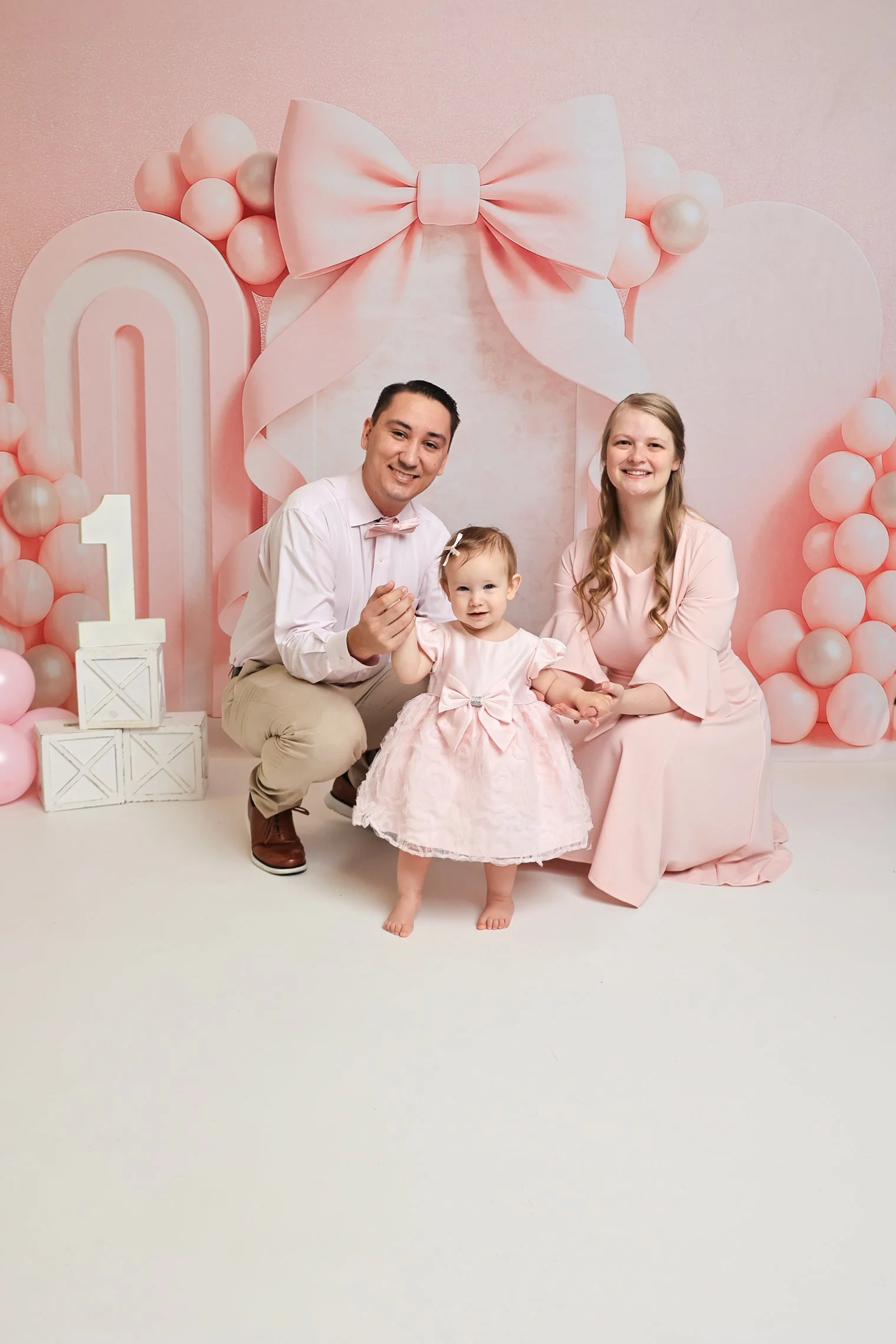 Family celebrating a first birthday with pink decorations, balloons, and a large pink bow backdrop.